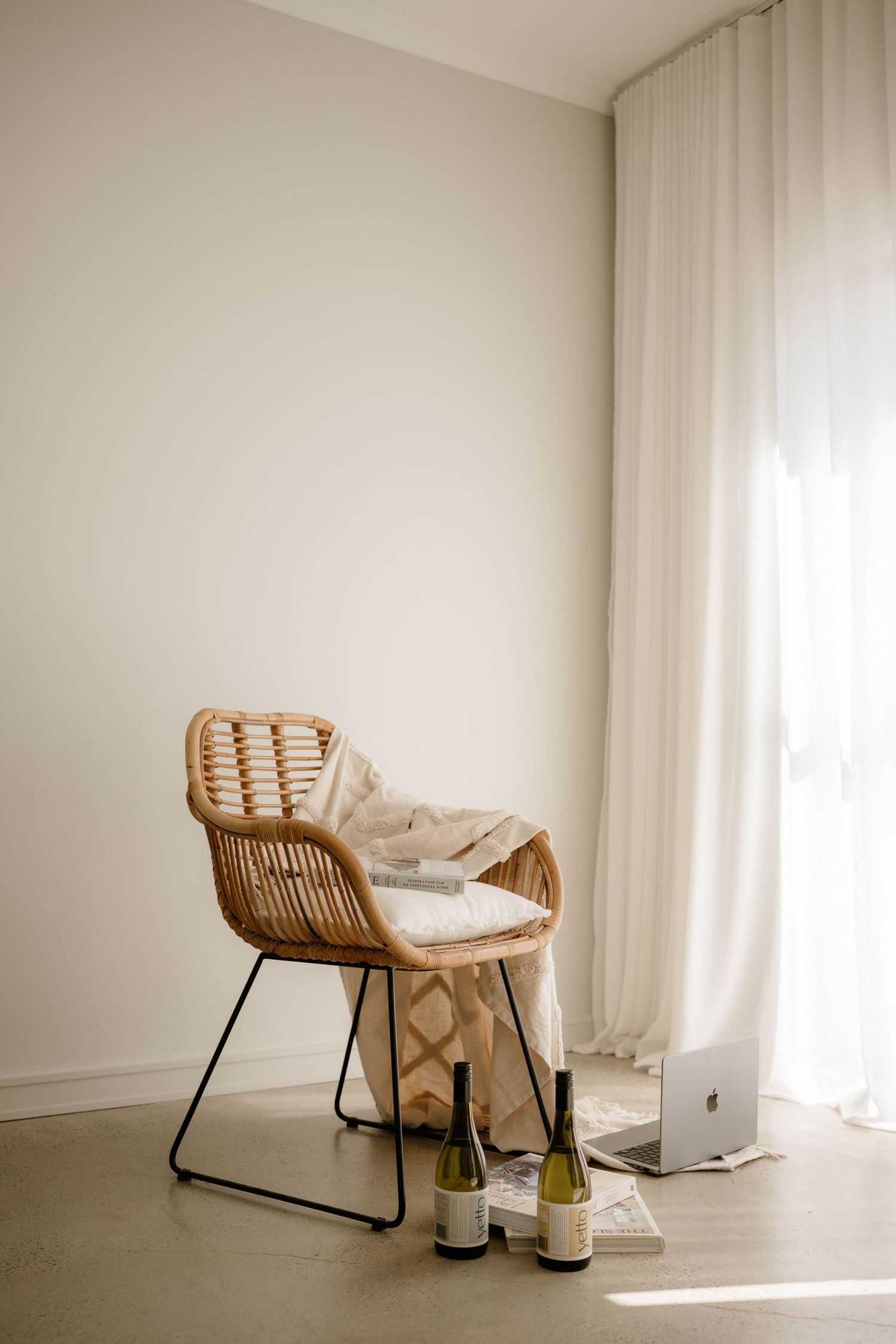 Modern rattan chair with blanket beside white curtains and two wine bottles on floor.