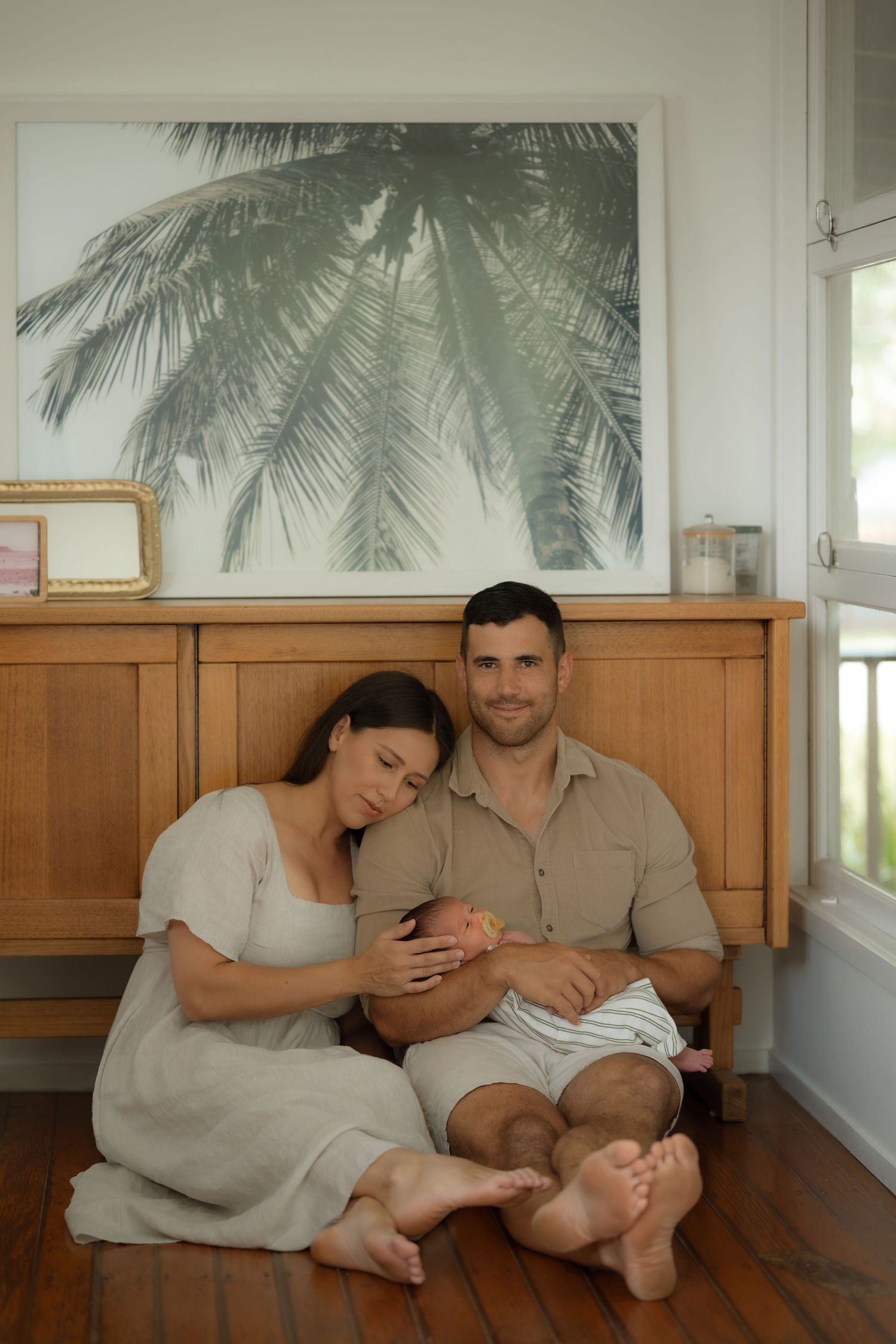 Parents sitting on floor cradling newborn baby beneath palm tree wall art.