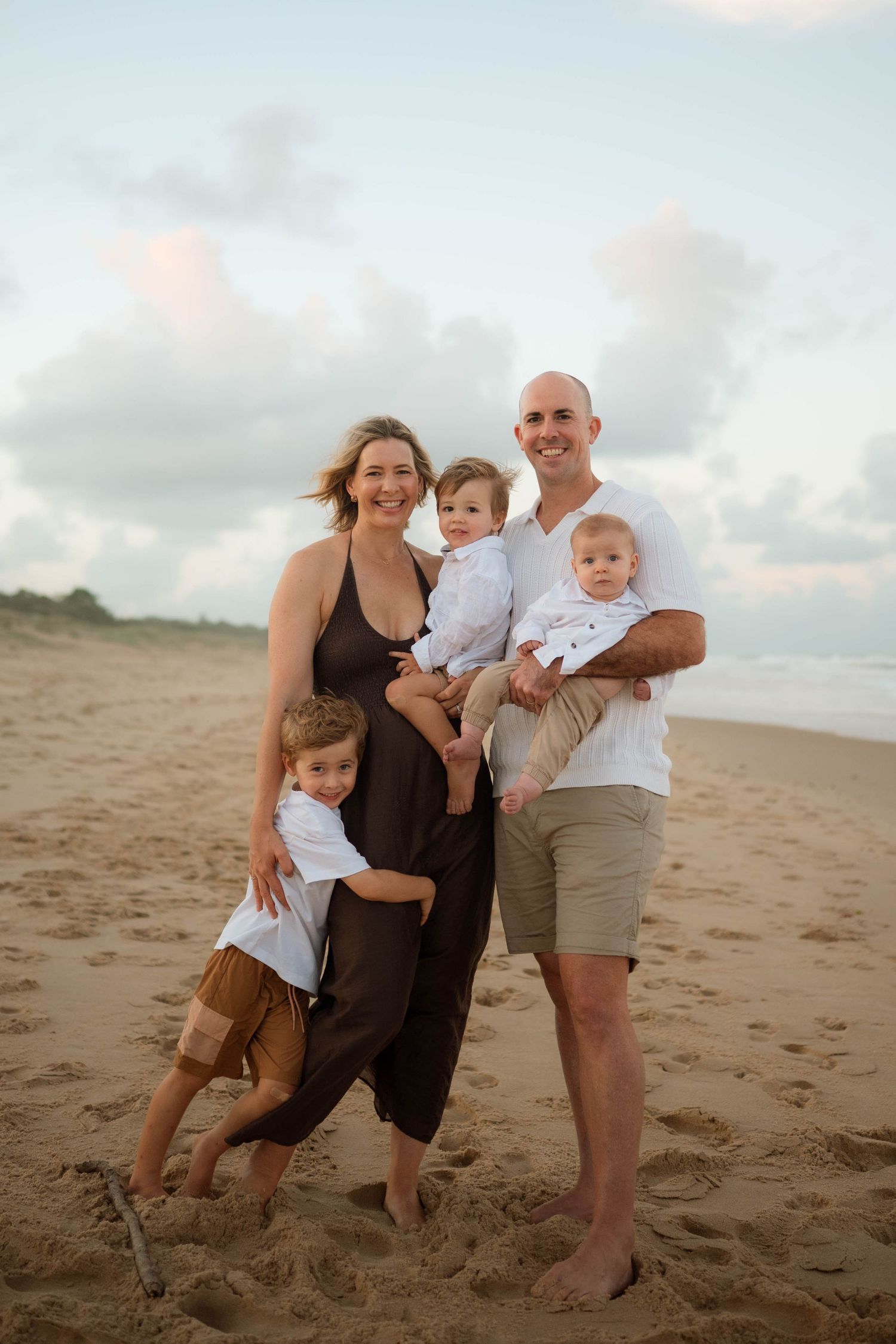 Happy family of five posing on a sandy beach at sunset, parents holding toddlers with young boy leaning against mom.