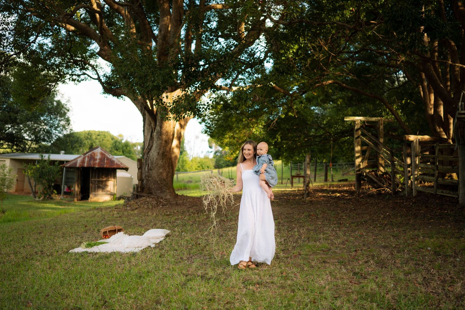 Mother in white dress lifting laughing baby outdoors under large trees on a sunny farm property.