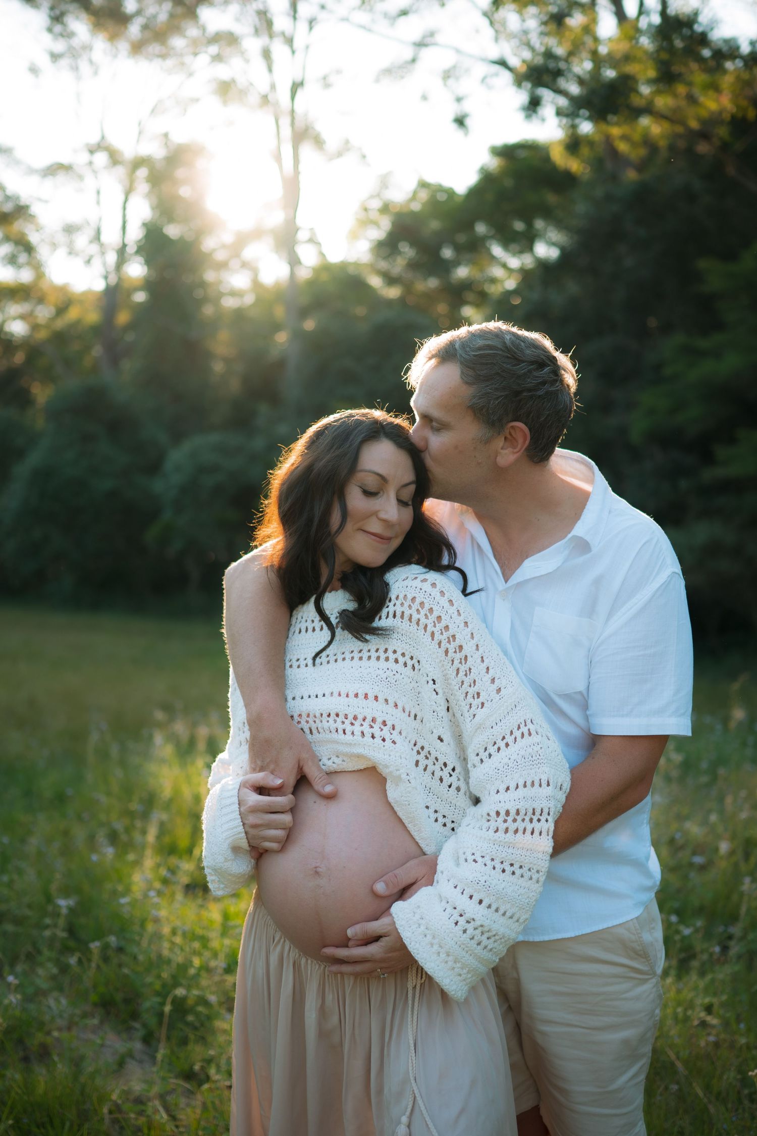 Couple sharing a tender moment in a sunlit field, man kissing pregnant womans head while holding her bump.