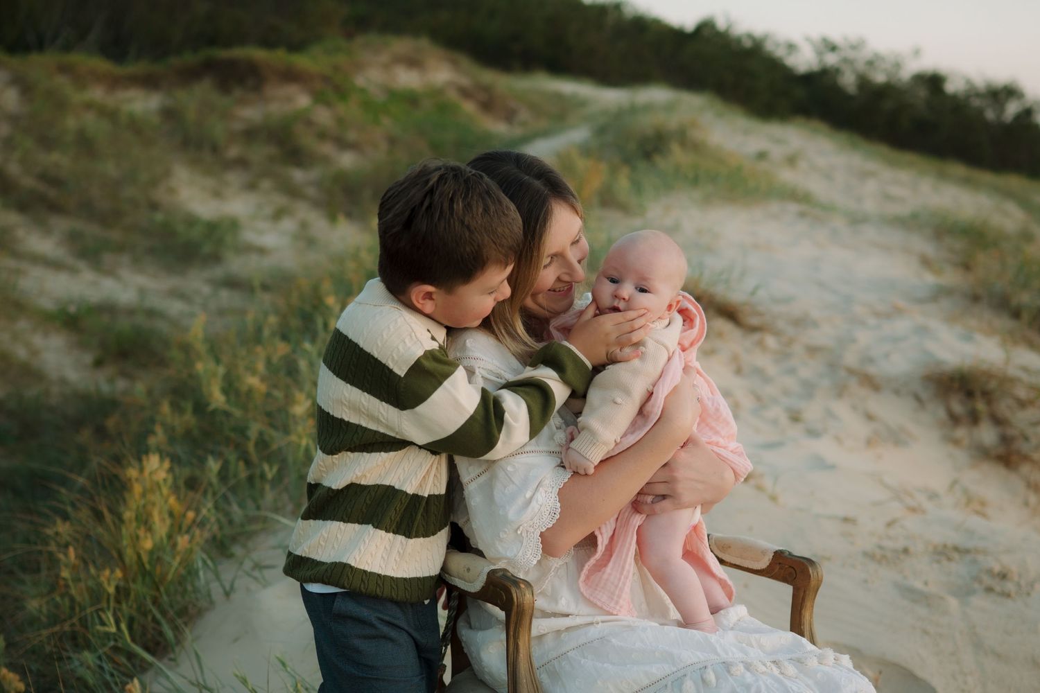 Mother sitting on chair at beach with toddler son kissing her cheek while she holds newborn baby in pink outfit.