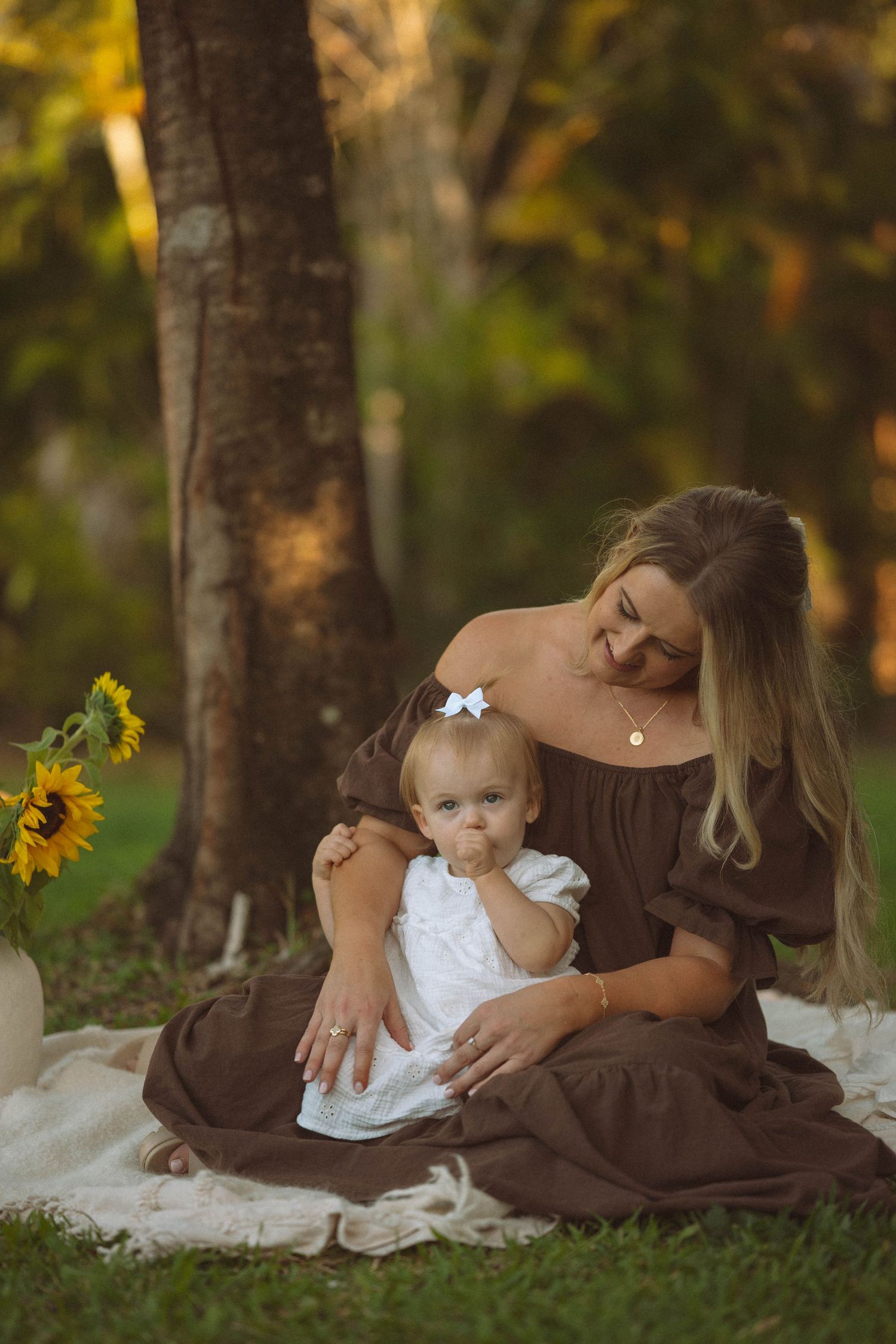 Blonde mother in brown dress sitting outdoors with baby girl near sunflowers at golden hour.