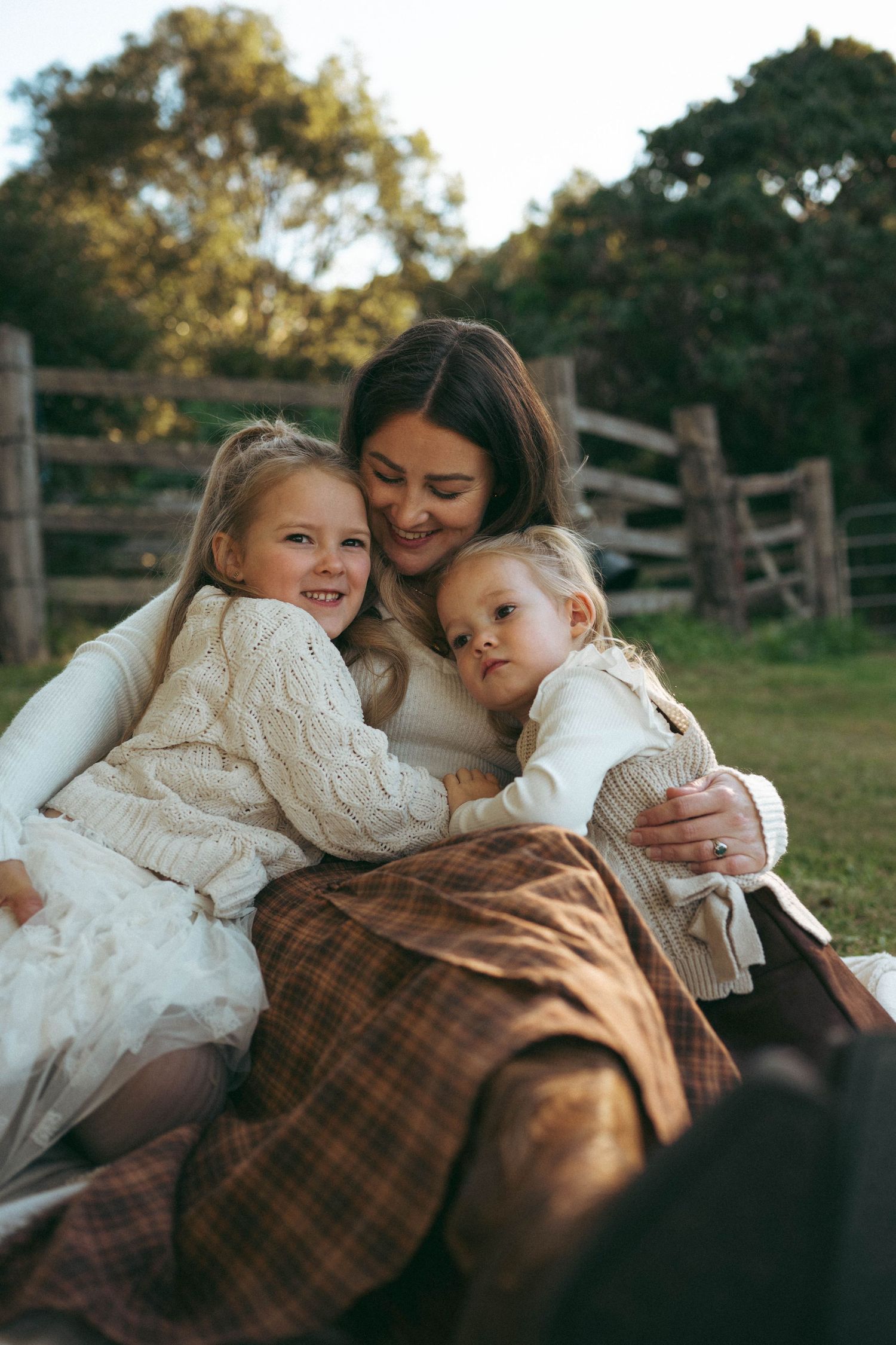 A smiling mother sits on a blanket outdoors embracing her two young daughters in cozy knit sweaters near a wooden fence.