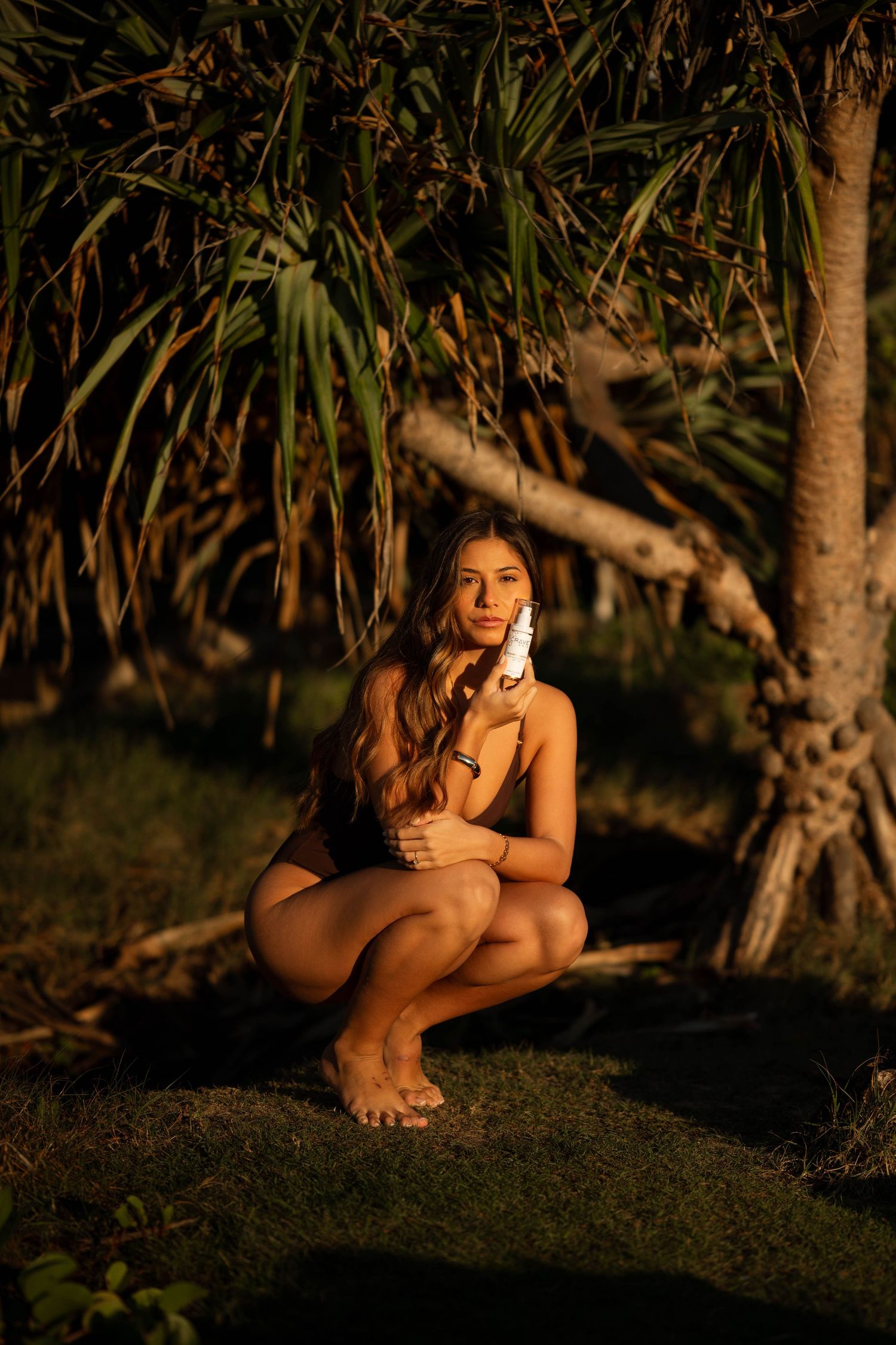 Woman in bikini crouching near tropical palm trees in golden sunset light, holding a white object.