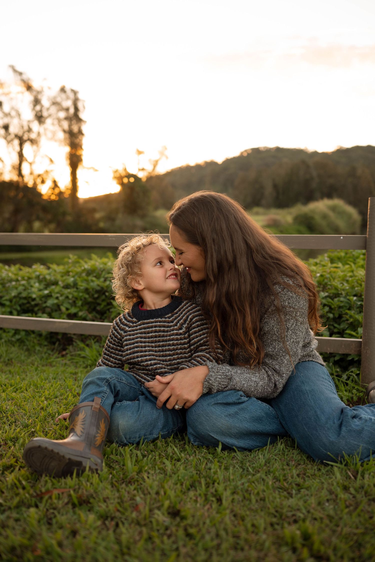 A mother and young child share a tender moment sitting on grass at golden hour near a wooden fence.