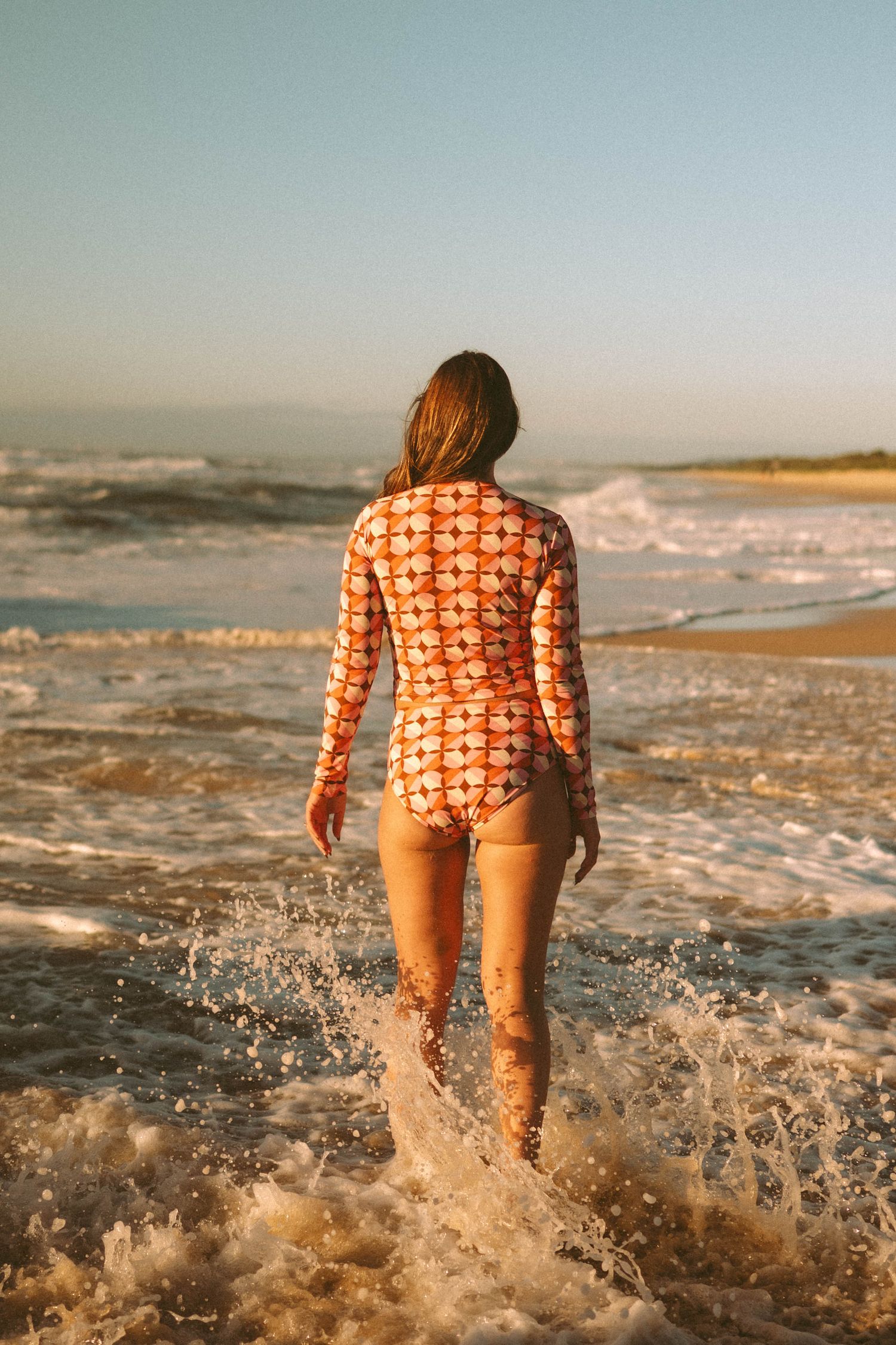 Back view of woman in orange floral long-sleeve one-piece swimsuit walking into ocean waves.