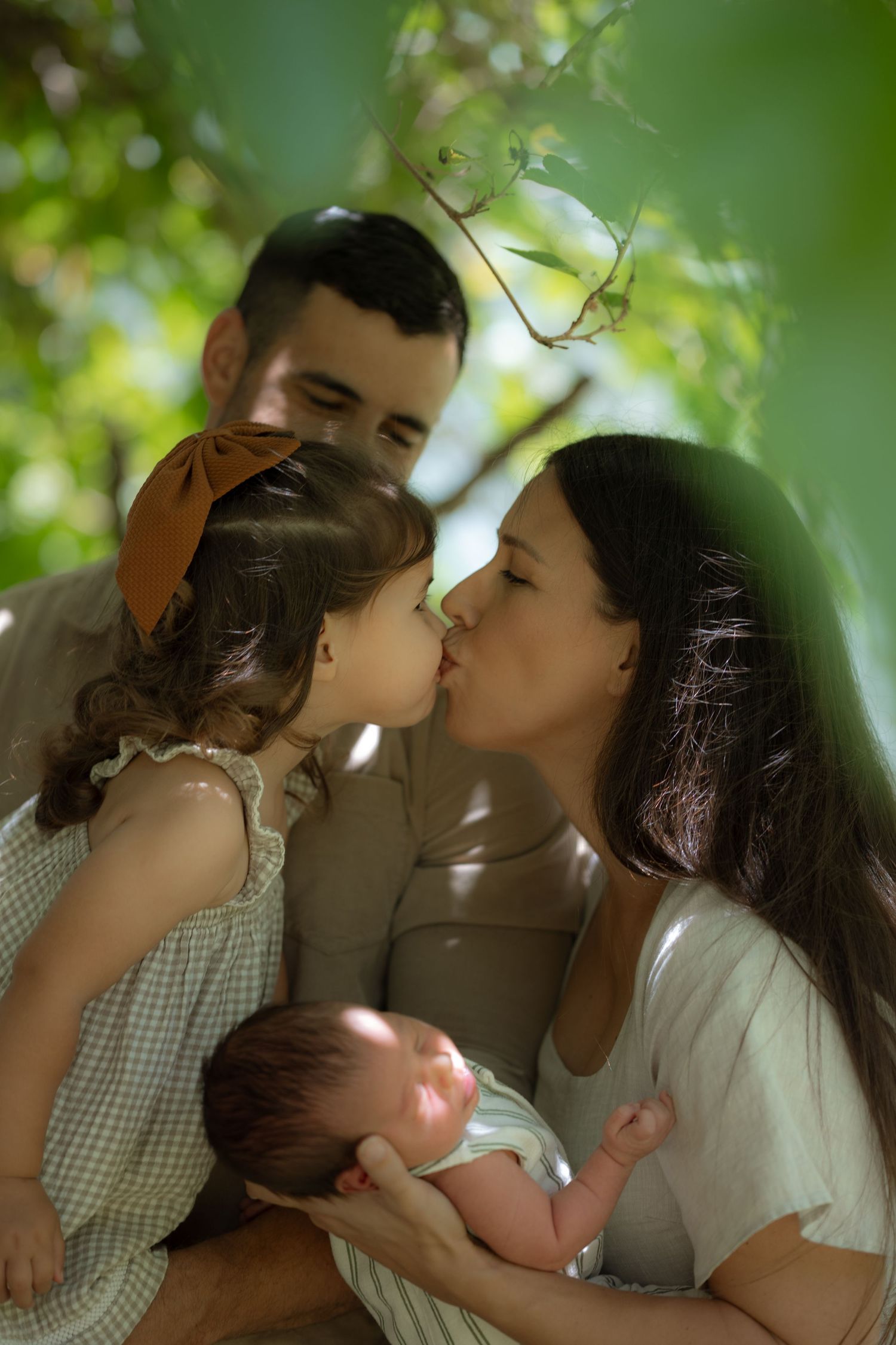 Mother kissing toddler daughter while father holds newborn baby in dappled sunlight under green trees.