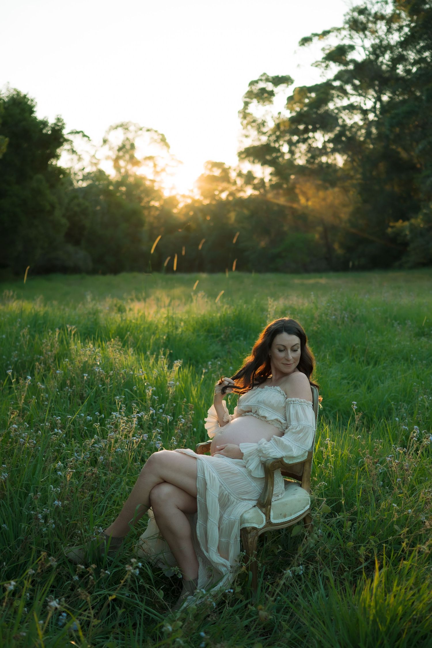 Serene pregnant woman in white lace dress sits in vintage chair in sunlit green meadow.