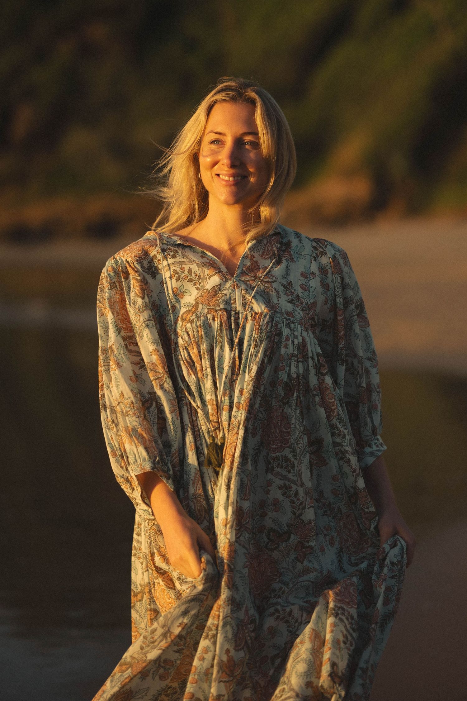 Smiling blonde woman in floral maxi dress standing on a beach at golden hour sunset.