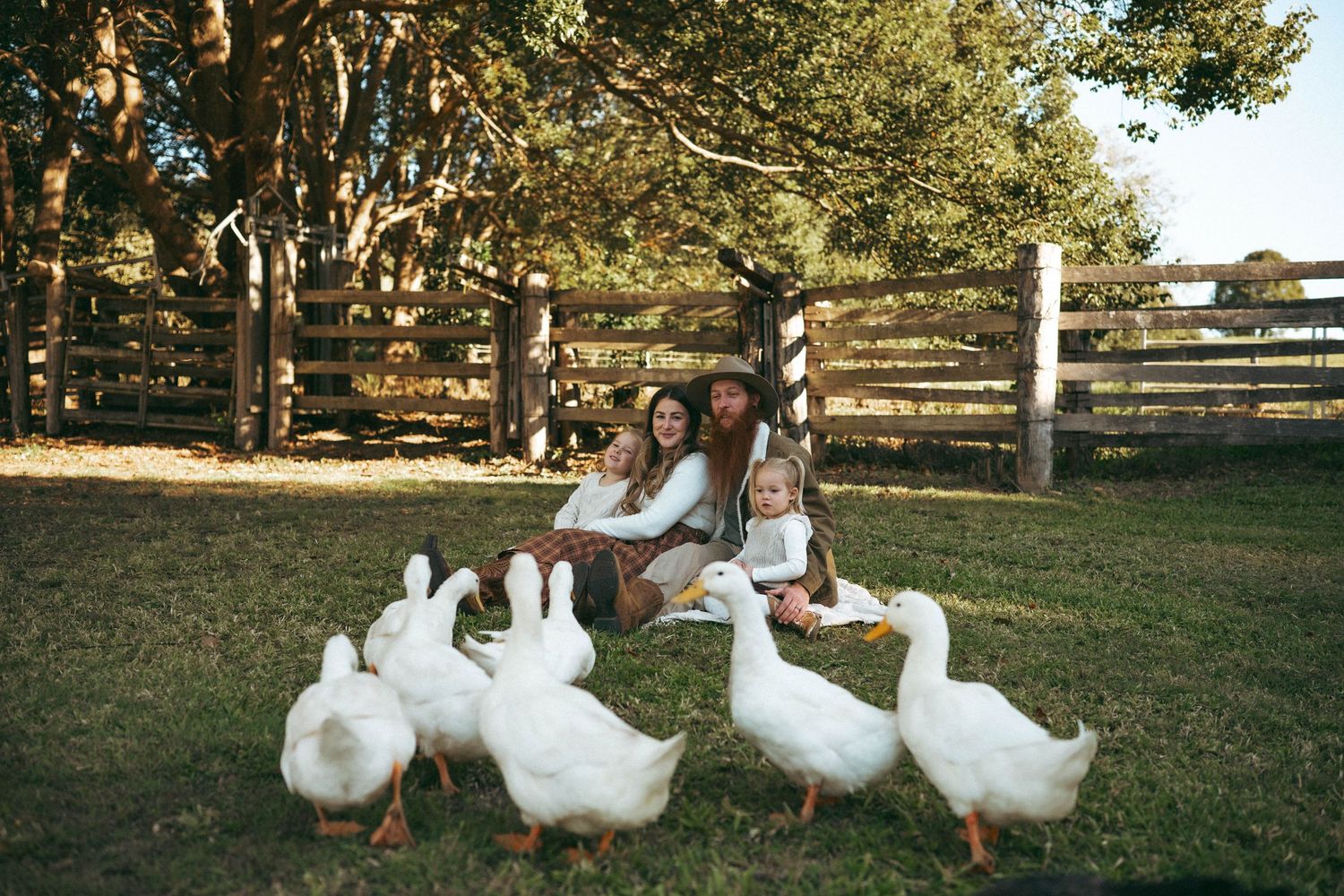 Family with two children sitting on grass surrounded by white geese near a wooden fence on a sunny farm.