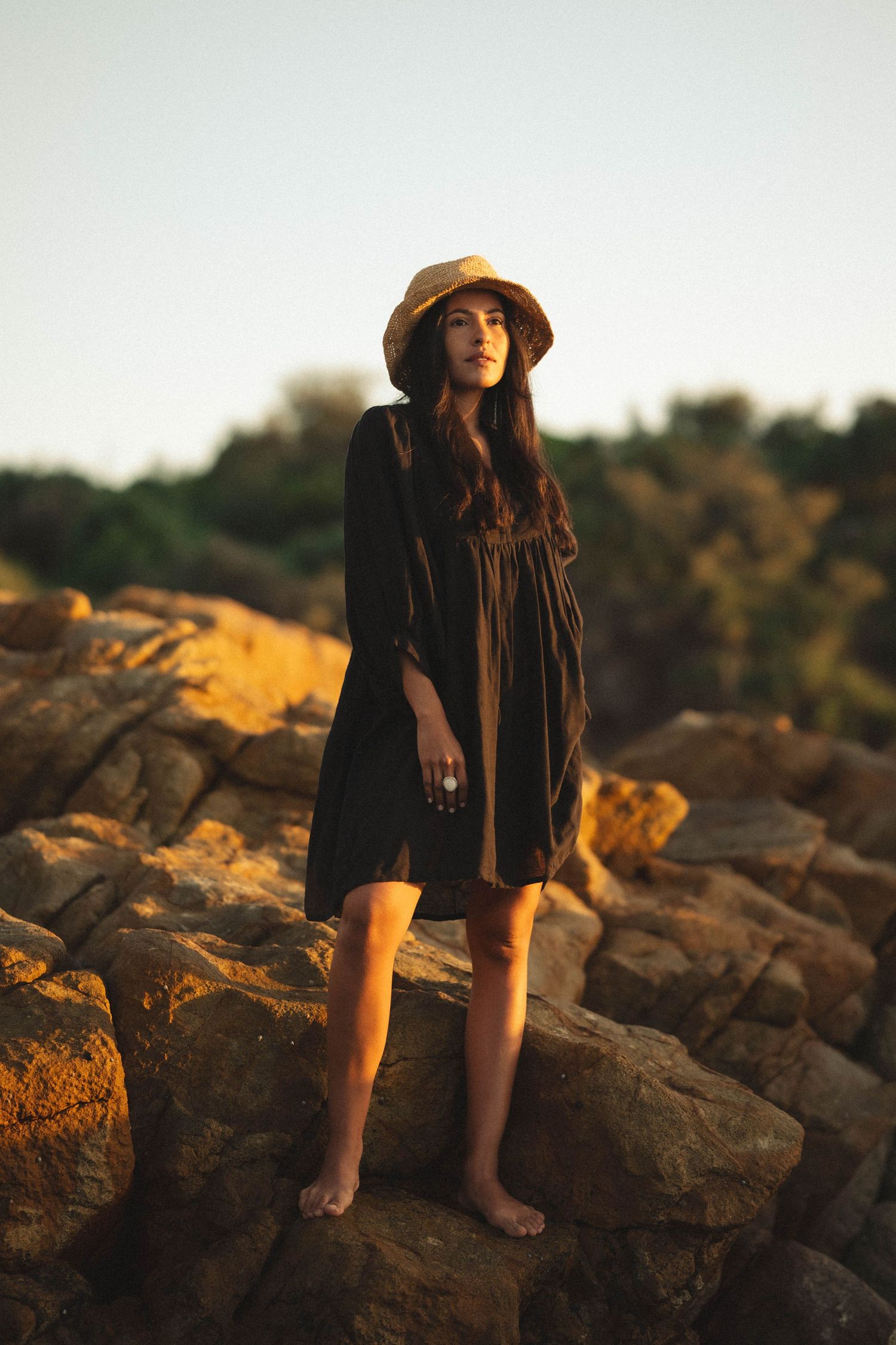 Barefoot woman in black dress and straw hat standing on golden rocks at sunset.