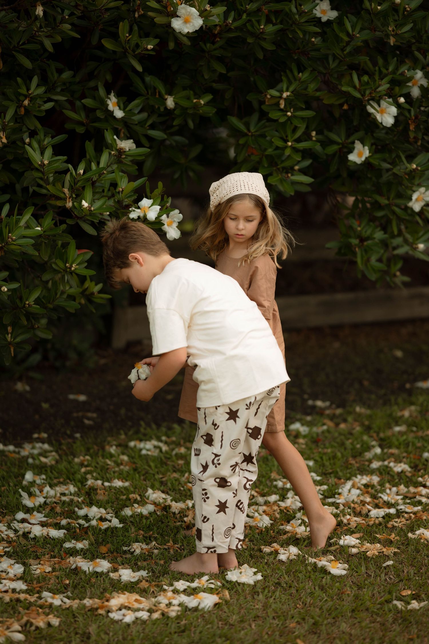 Two children picking up white flowers from grass beneath a flowering shrub outdoors.