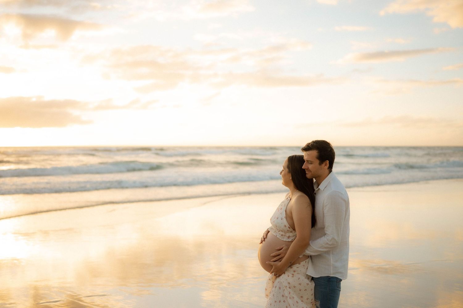 Expectant couple kissing on reflective wet sand beach at golden sunset with soft ocean waves behind.
