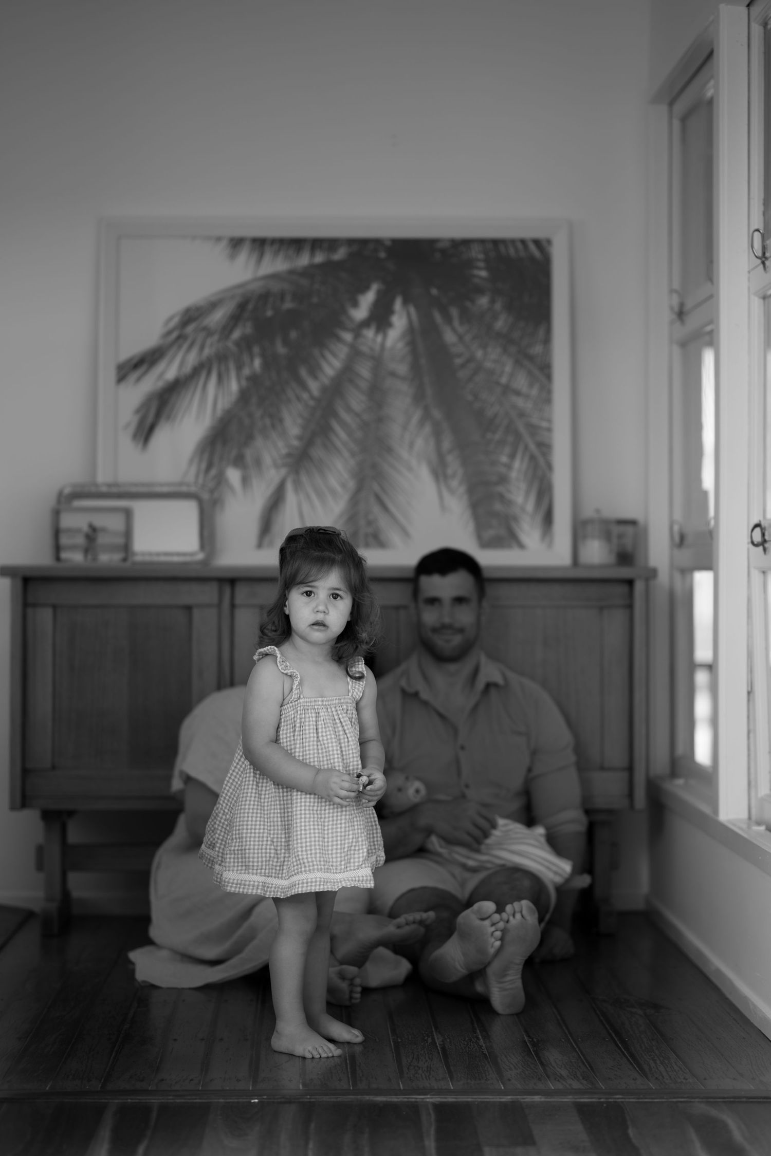 Black and white portrait of toddler girl standing with father holding newborn indoors.
