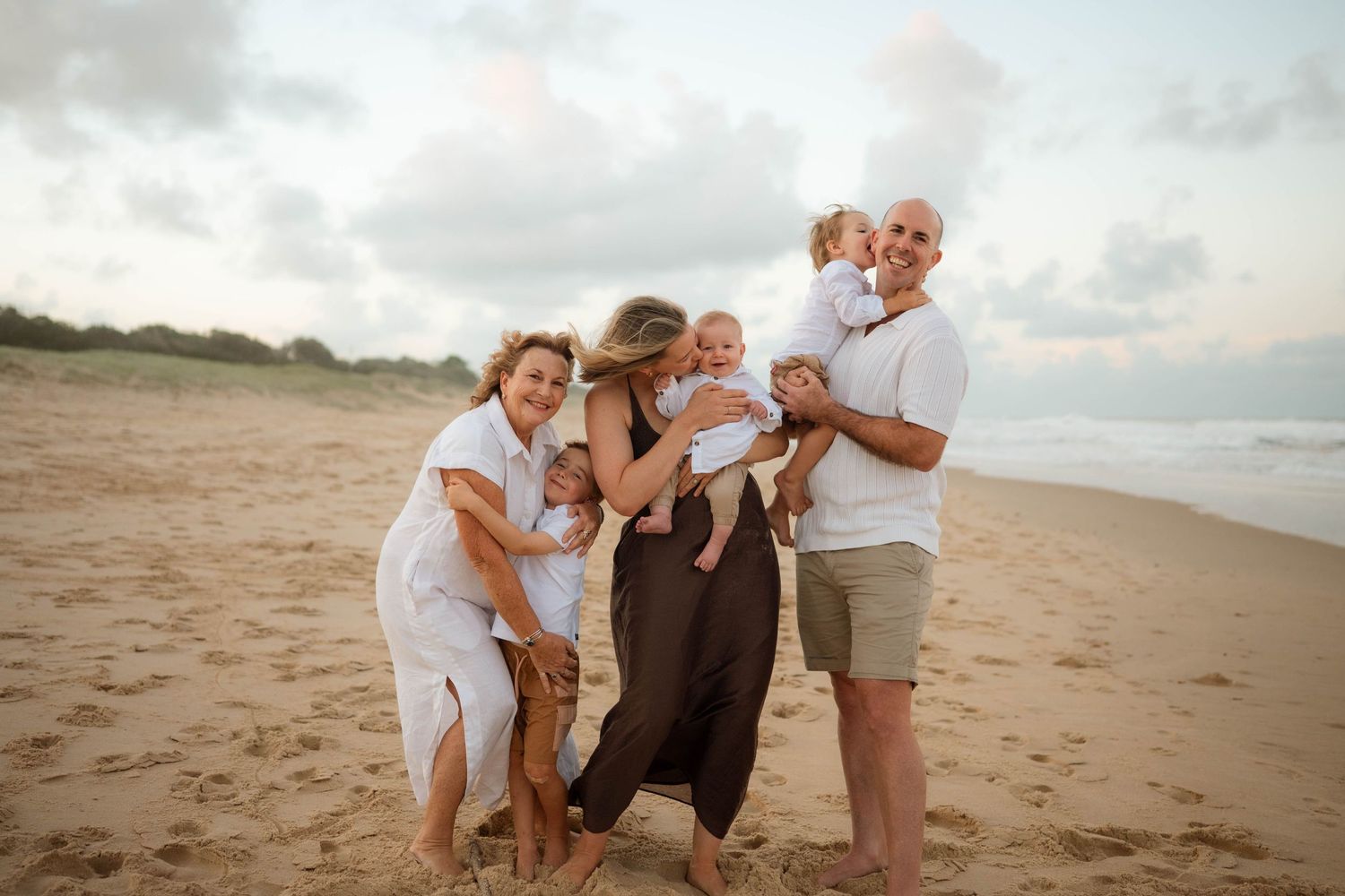 Joyful extended family embracing and laughing together on a sandy beach during golden hour sunset.