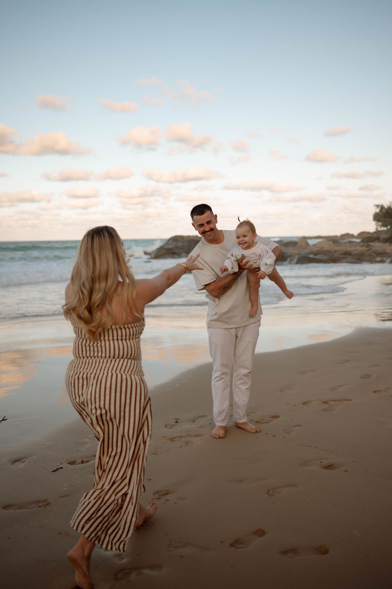 Family enjoying a beach sunset, mother reaching toward father holding baby, waves and rocks in background.