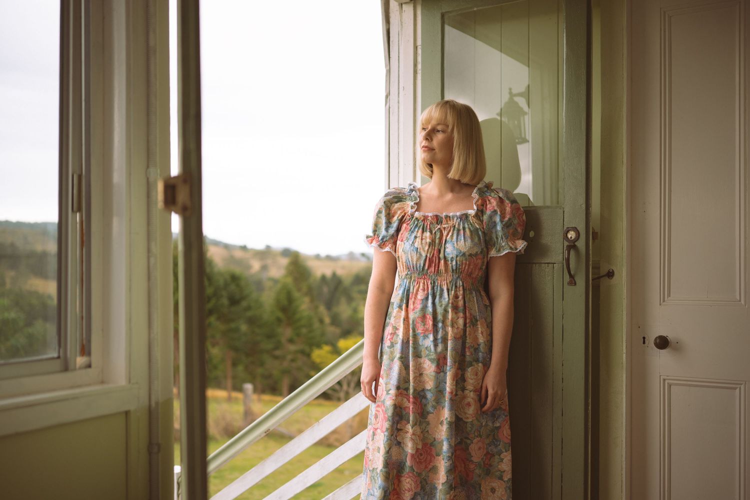 Woman in floral dress standing by open door with scenic countryside view.