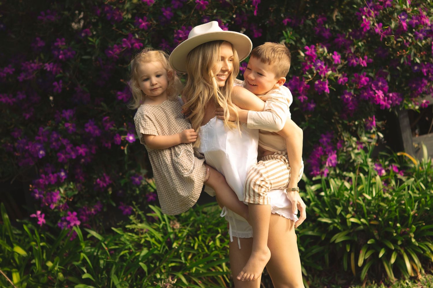 Woman in white hat holding two young children outdoors with purple flowering bushes in background.