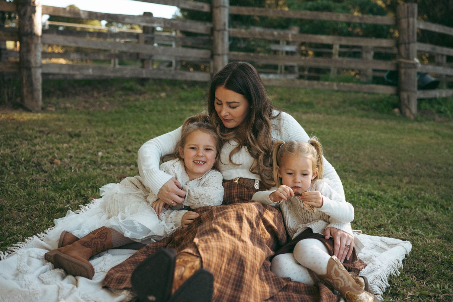 A mother sits on a plaid blanket outdoors with two young children, all wearing cozy cream sweaters in a fall setting.