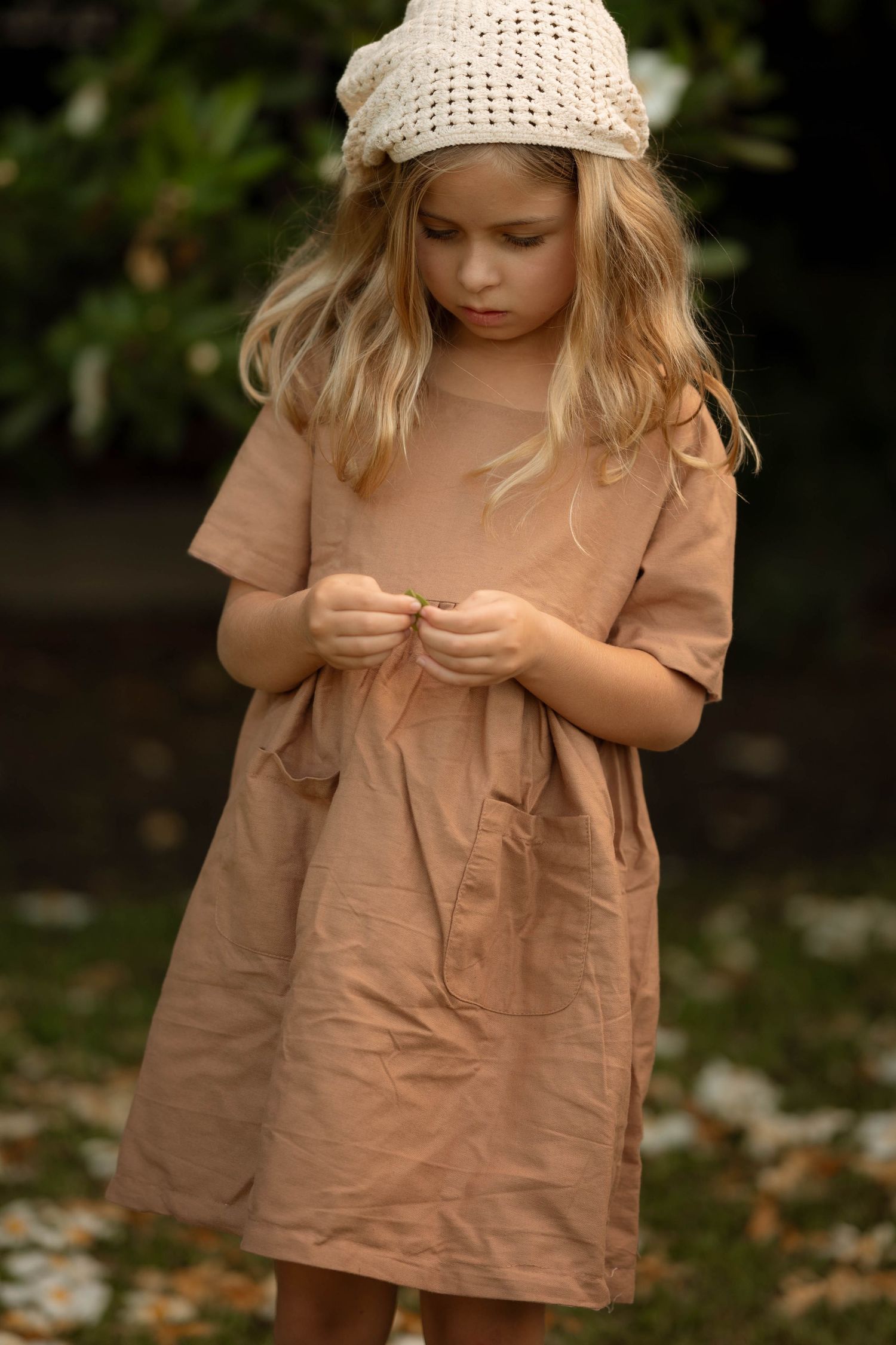 Young girl in a dusty rose linen dress and cream crochet hat, looking down outdoors.