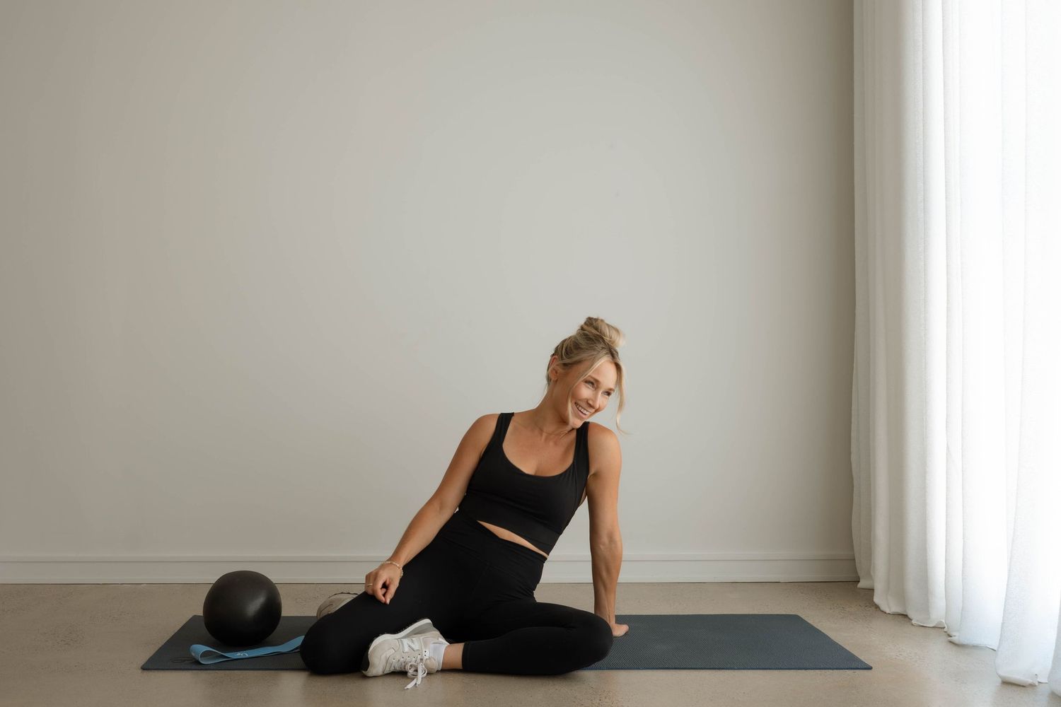 Smiling woman in black workout attire sitting on yoga mat with exercise ball nearby.