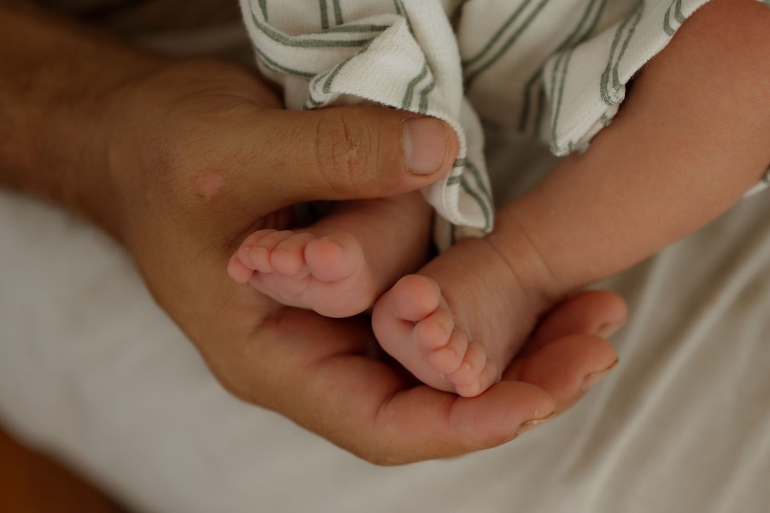 Close-up of adult hands gently cradling tiny newborn baby feet in soft light.