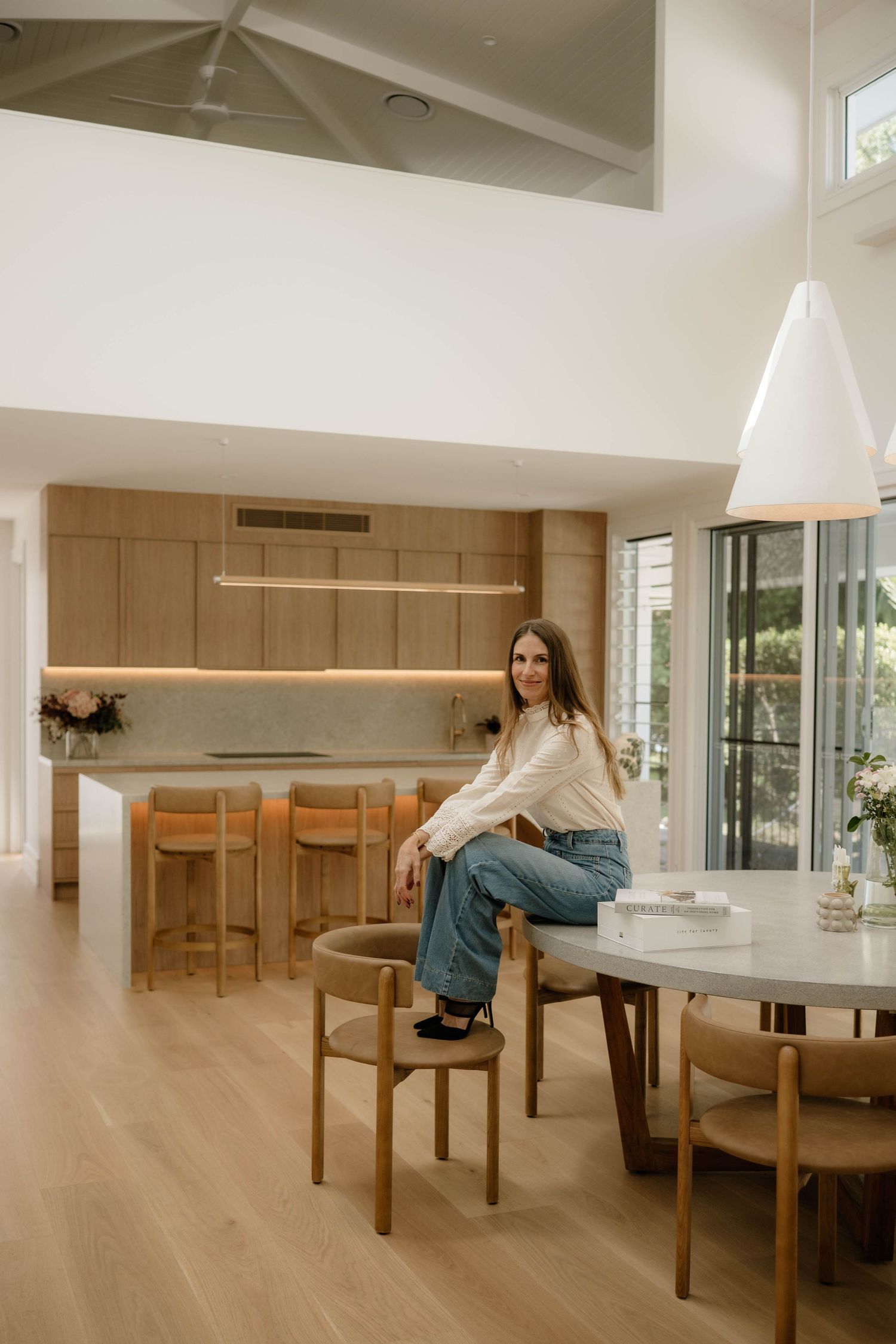 Woman in white blouse and jeans sitting on dining table in modern open-plan kitchen.