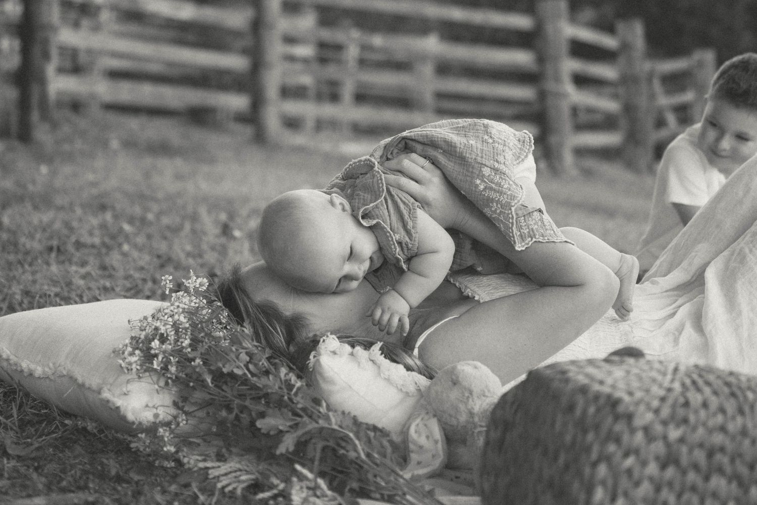 Black and white photo of a mother lying on a blanket in a garden, joyfully lifting her baby overhead.