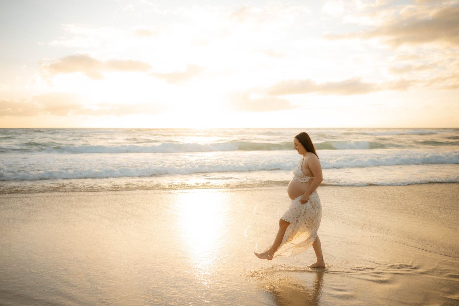Pregnant woman in white dress walking along a golden sunset beach with waves gently lapping the shore.