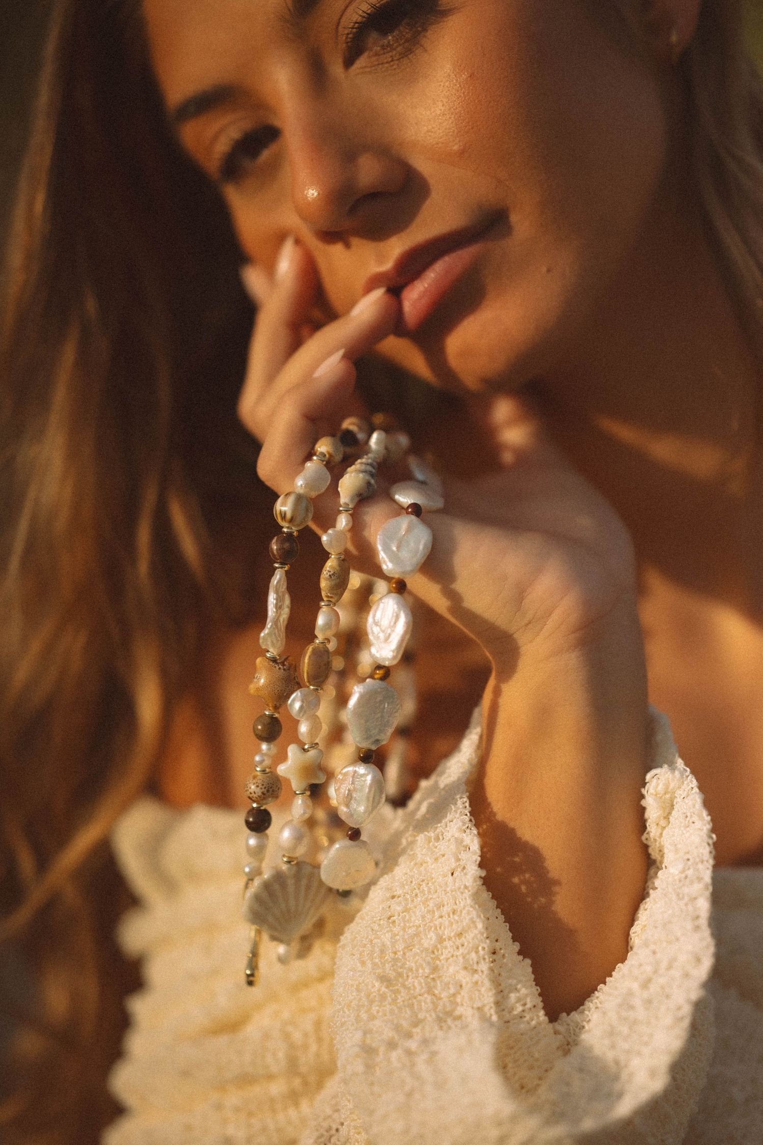 Woman holds layered pearl and shell necklaces in warm golden sunset light.