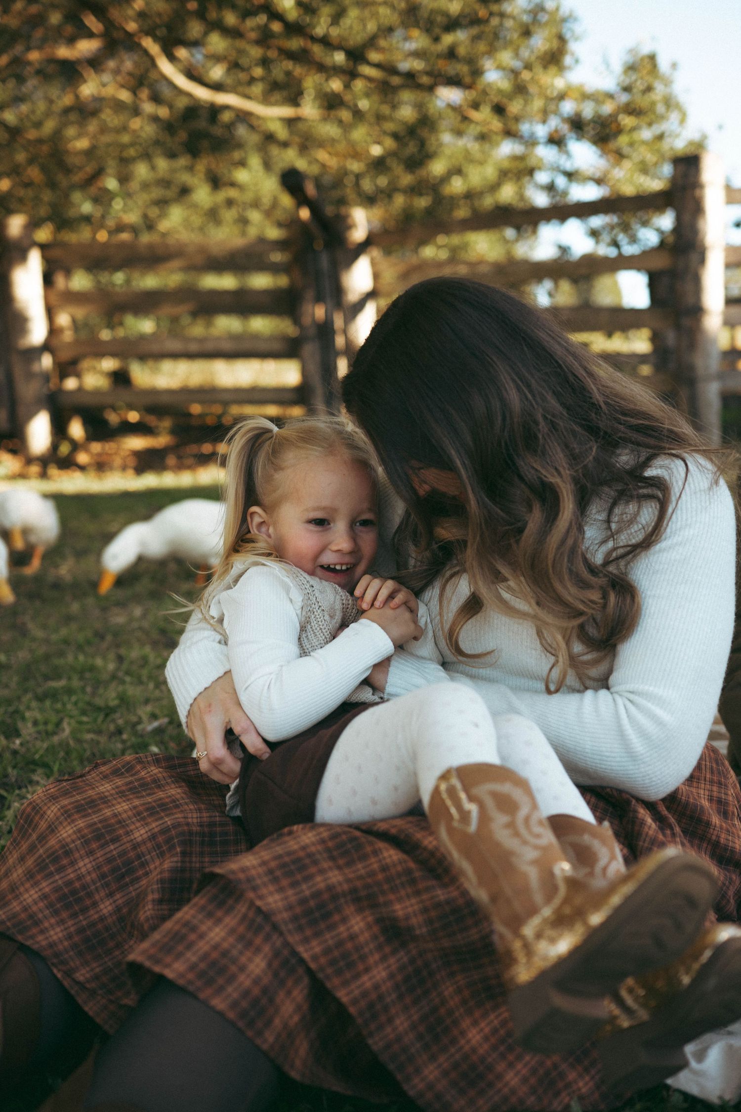 Mother laughing with toddler daughter on plaid blanket at farm, wearing white tops and brown cowboy boots.