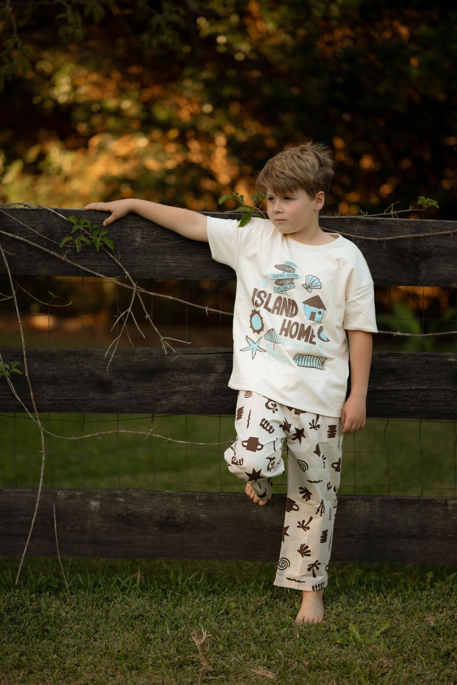 Boy in cream Island Home pajamas leaning against wooden fence outdoors, holding soccer ball, surrounded by green grass.