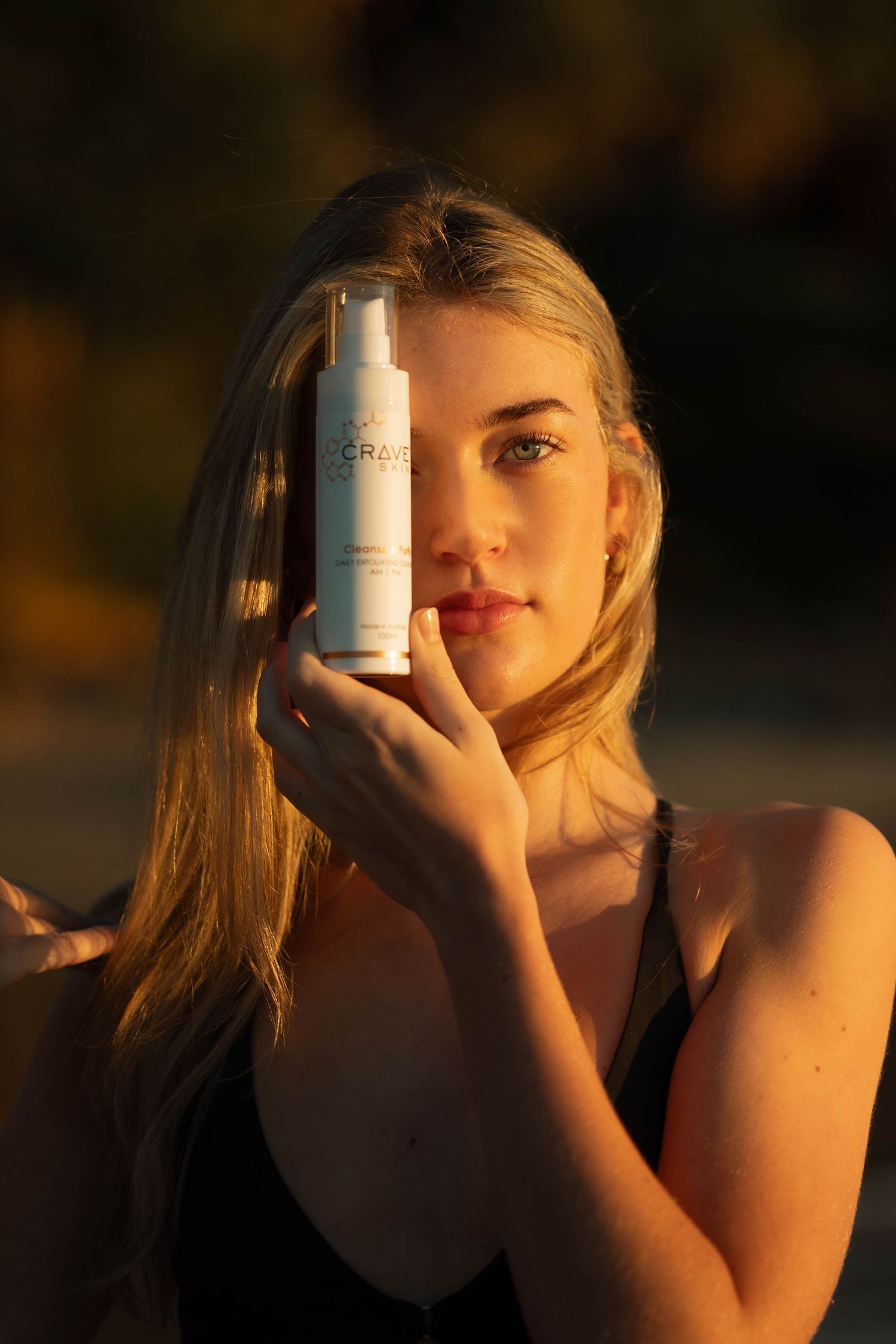 Blonde woman in black swimsuit holding white skincare product near face at golden hour.