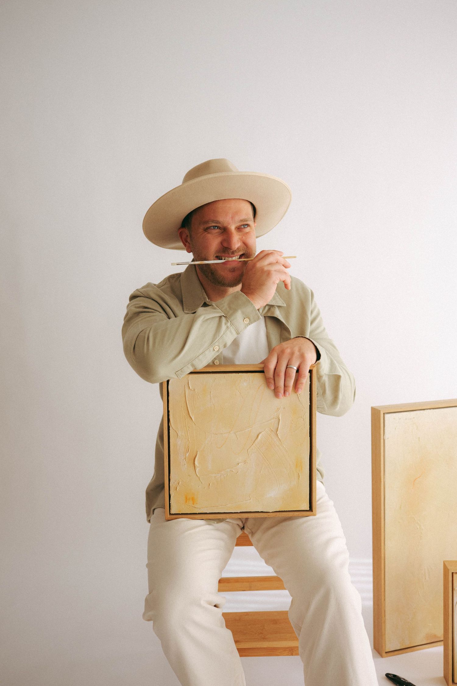 A smiling man in a wide-brim hat holds a canvas painting while biting a paintbrush in a bright studio.