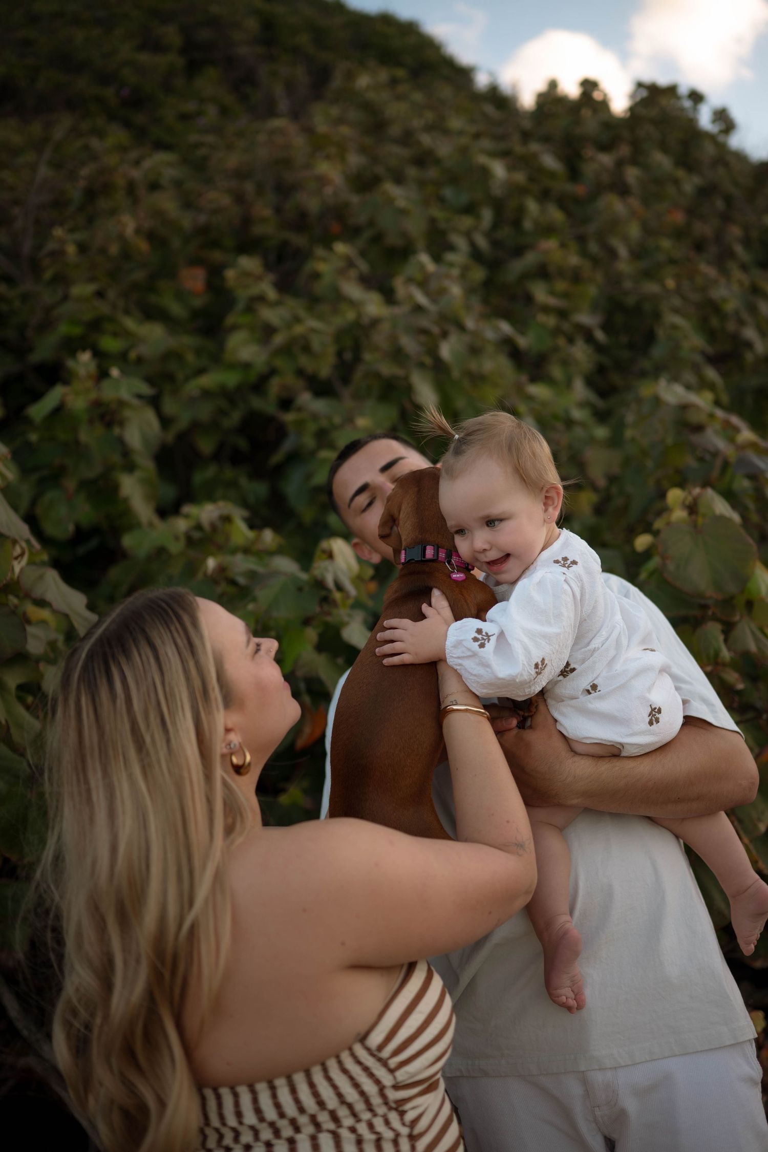 A happy family with a smiling baby in white dress, parents kissing child outdoors with lush green foliage background.