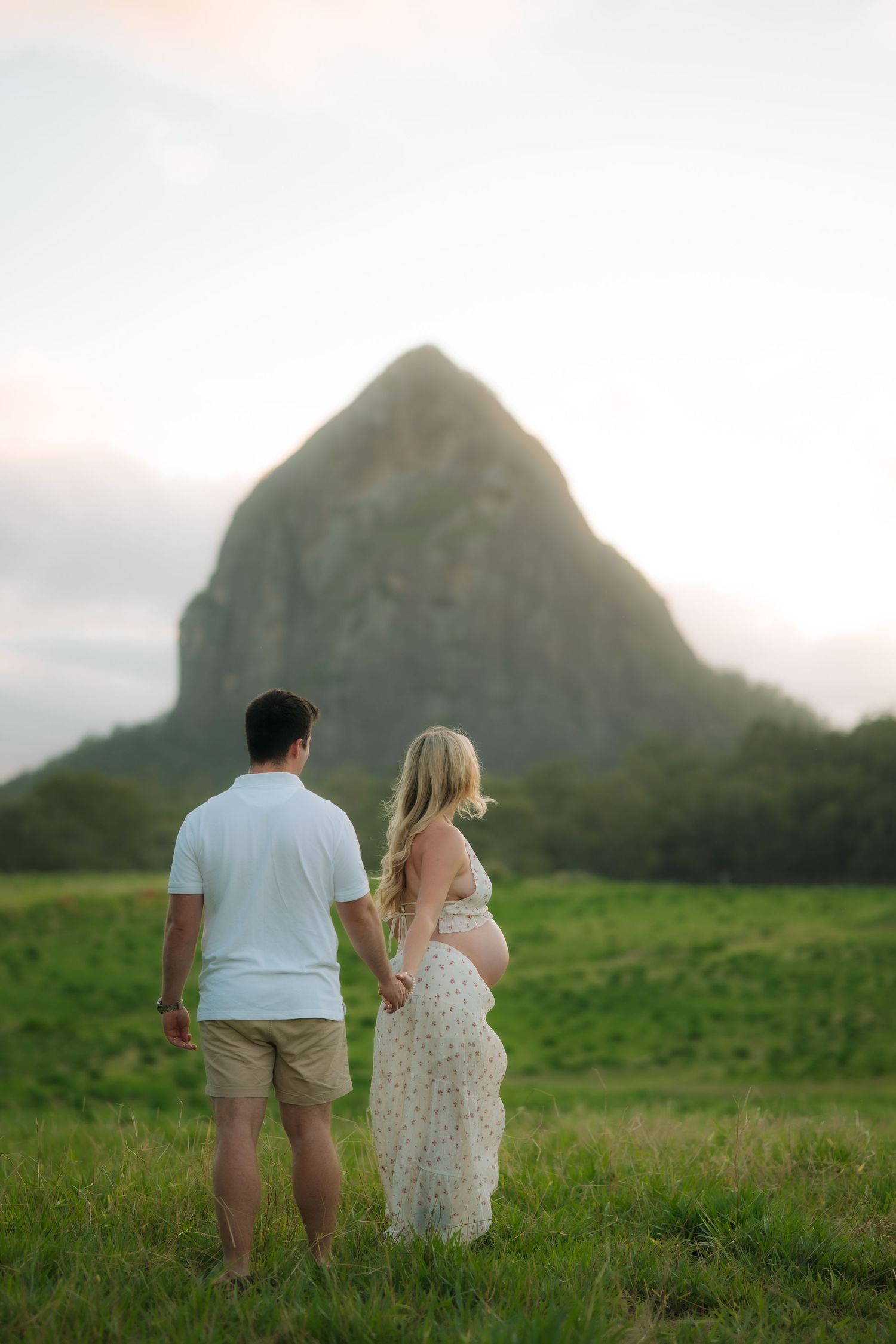 Expecting couple holds hands while facing a dramatic rocky peak in a green field during golden hour sunset.