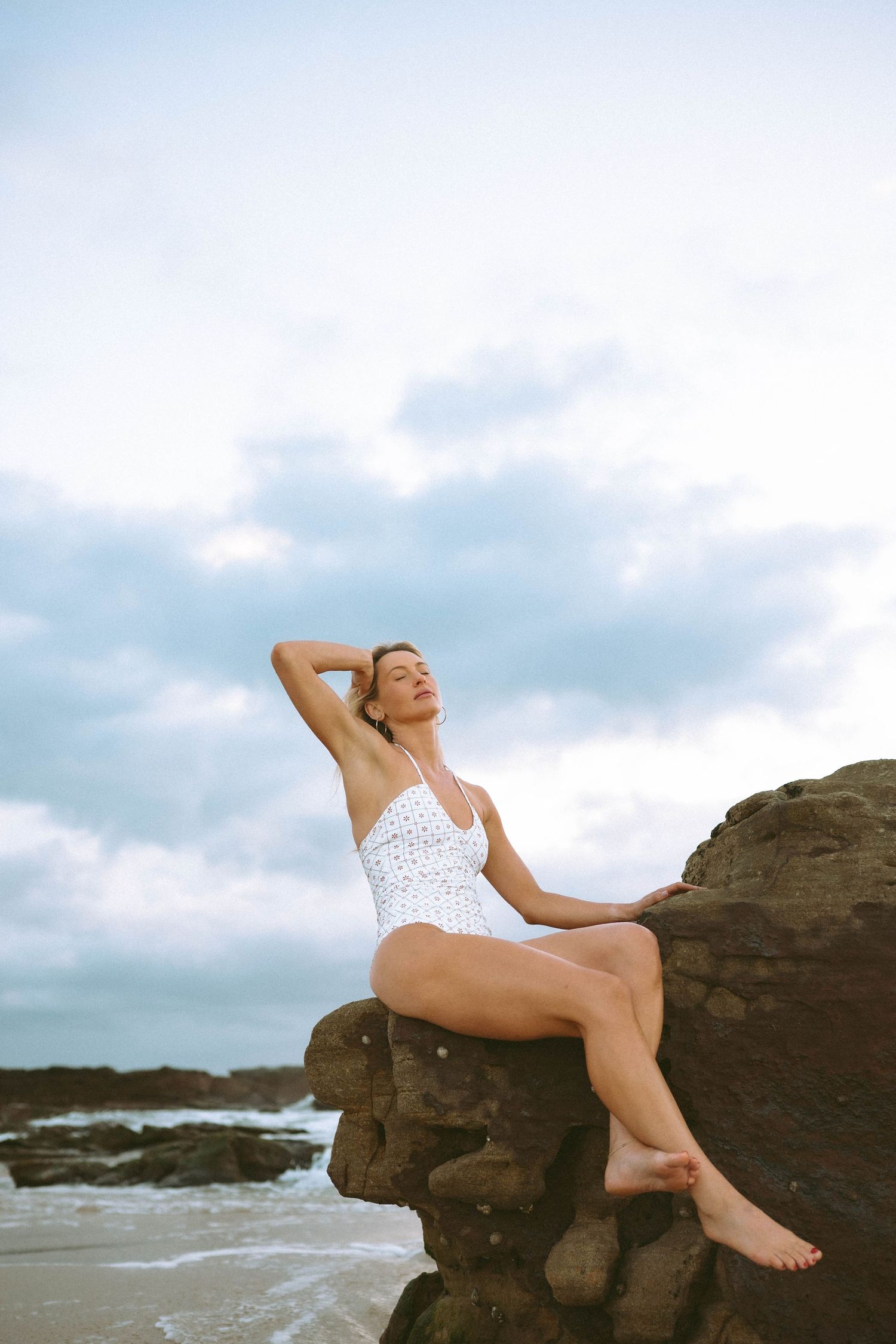 Woman in white one-piece swimsuit sitting on coastal rocks with eyes closed under dramatic cloudy sky.
