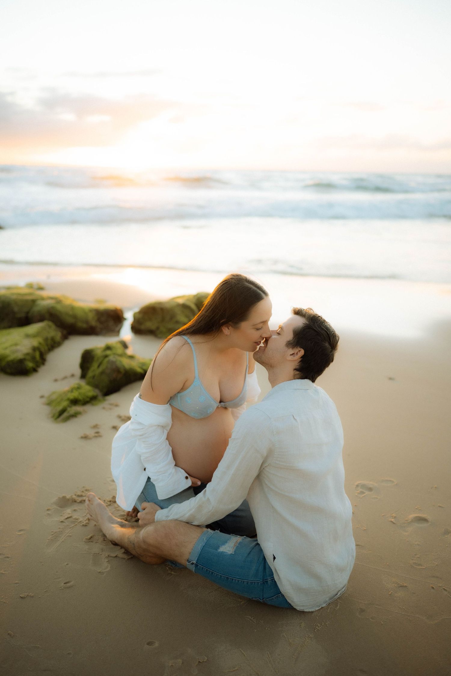 Couple kissing on sandy beach at sunset, man tenderly holding pregnant woman's belly.