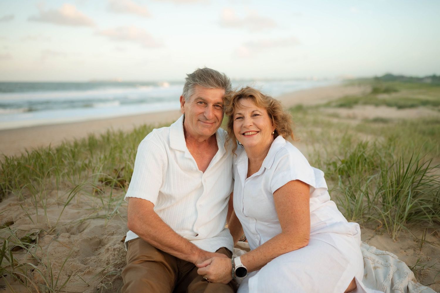 Smiling older couple sitting together on beach sand dunes at sunset.