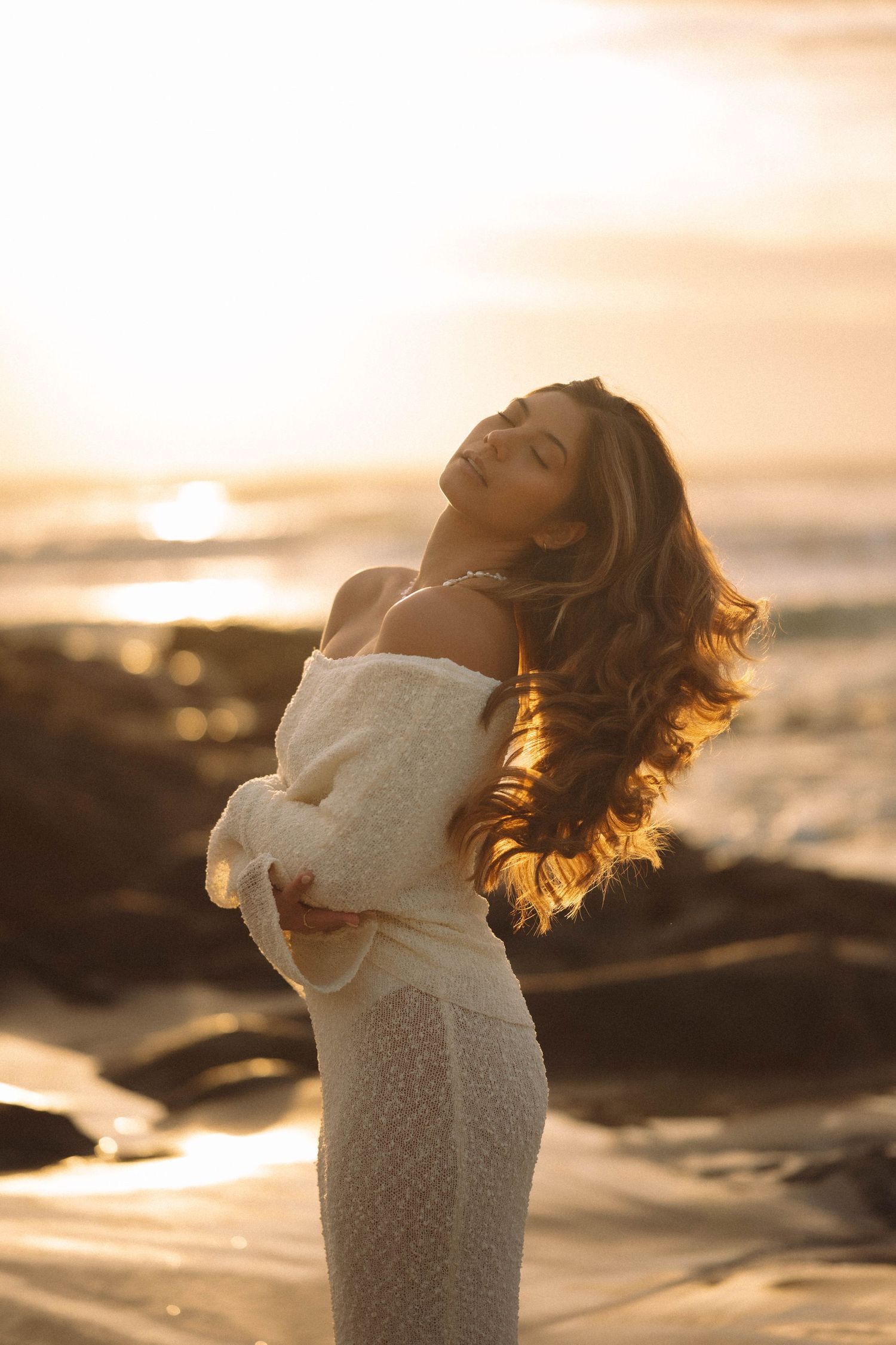 Woman in white off-shoulder lace dress poses gracefully on beach at golden sunset.