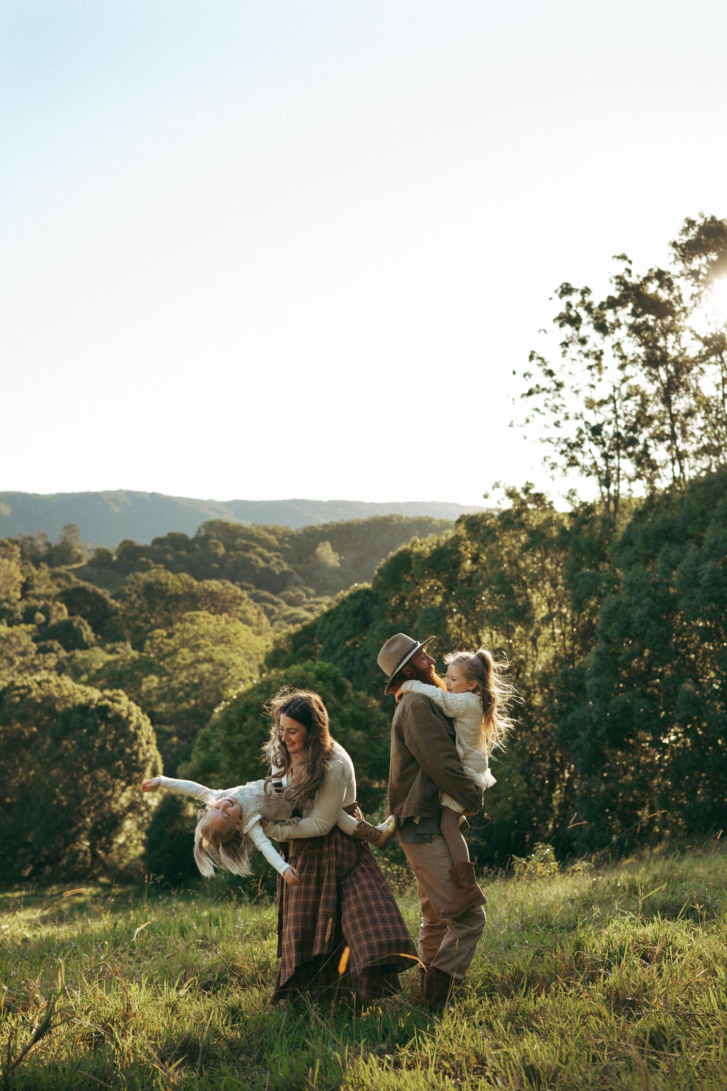 Family playing outdoors on a sunny hillside surrounded by lush green trees and rolling countryside landscape.