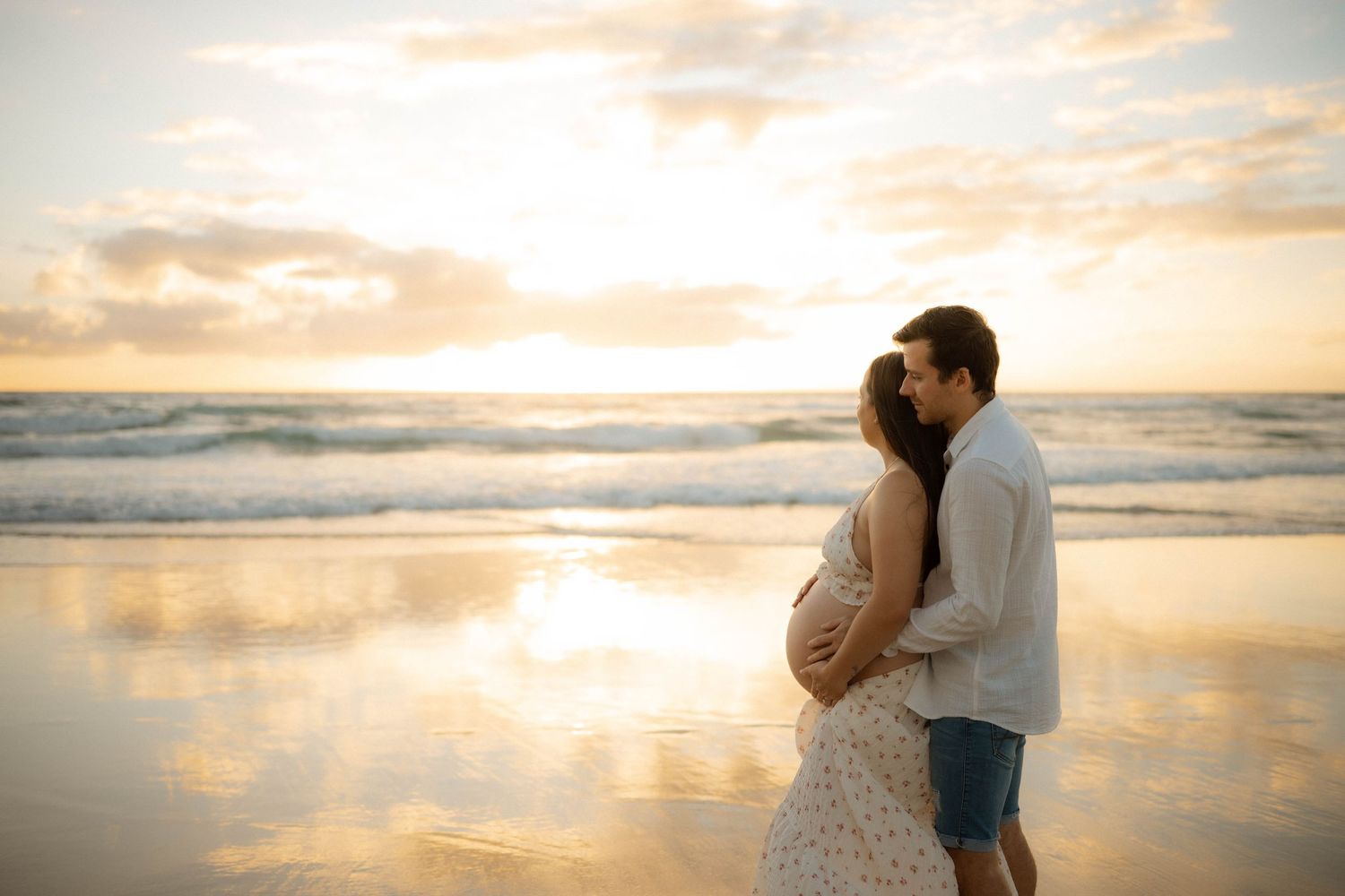 Couple sharing intimate kiss on beach at sunset, hands cradling pregnant belly in golden hour light.