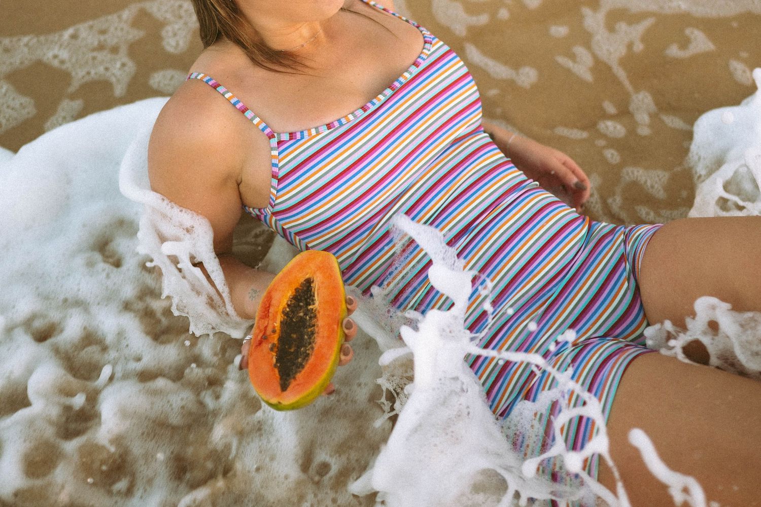 Woman in striped swimsuit sitting in ocean surf holding a halved papaya on sandy beach.