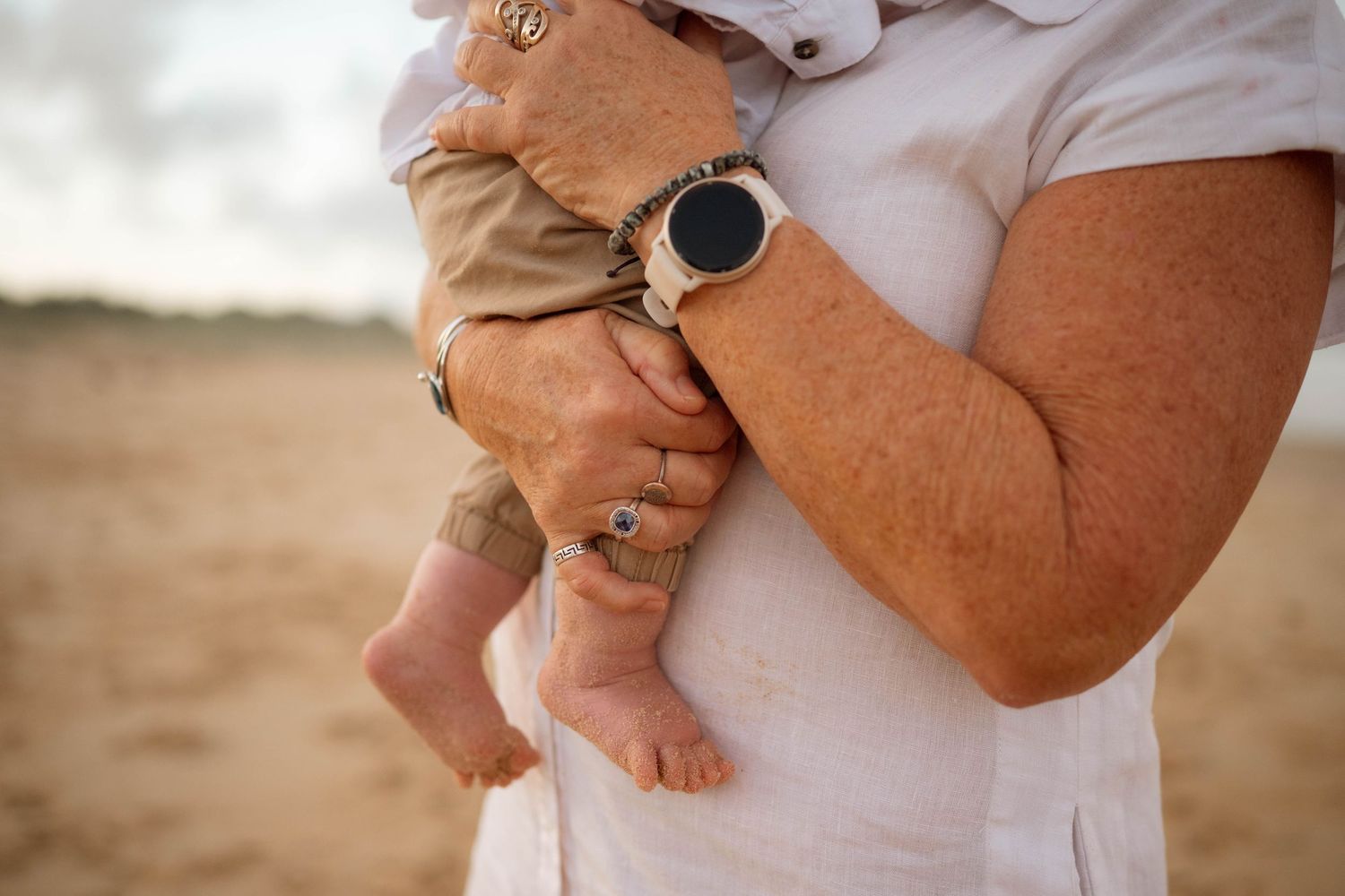 Close-up of grandparent holding a baby on the beach, showcasing a smartwatch and rings on sun-spotted hands.