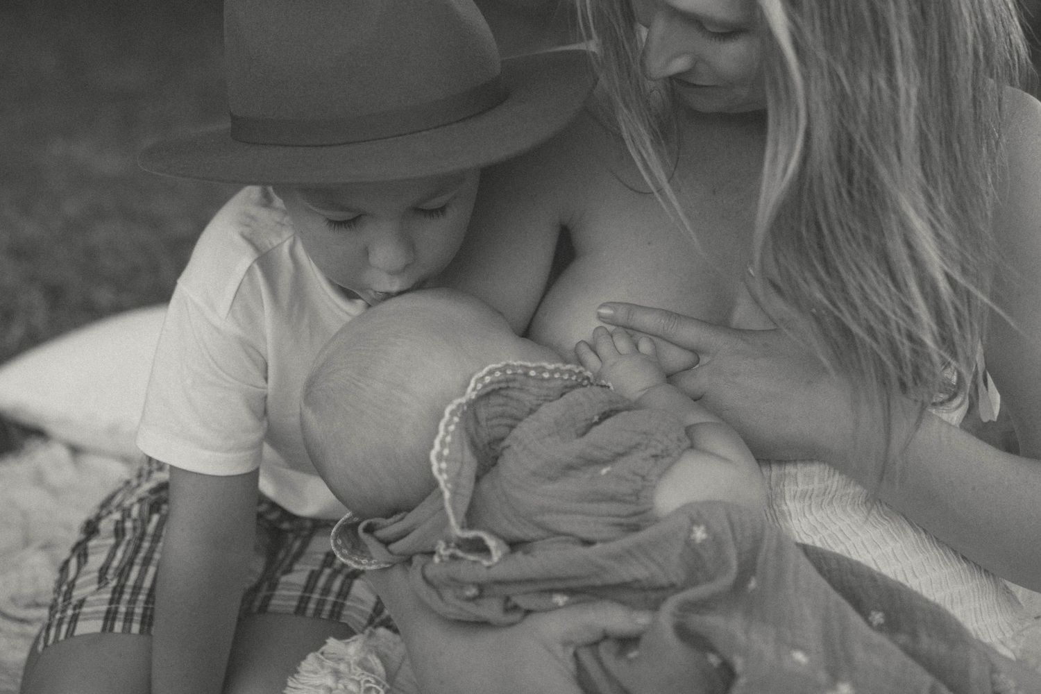 Black and white photo of a young child in a hat leaning in to kiss a newborn baby held by their mother.