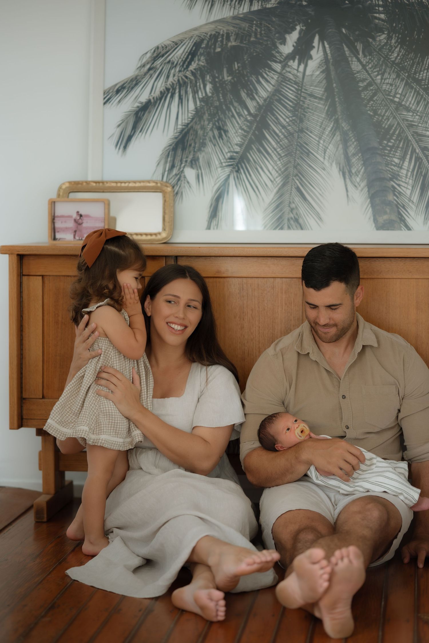Smiling family portrait with toddler and newborn baby sitting against wooden sideboard.