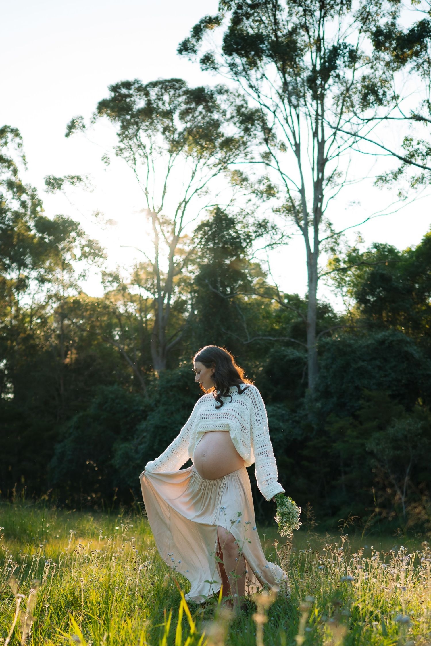 Pregnant woman in white knit sweater and flowing skirt walking through a sunlit meadow holding wildflowers.