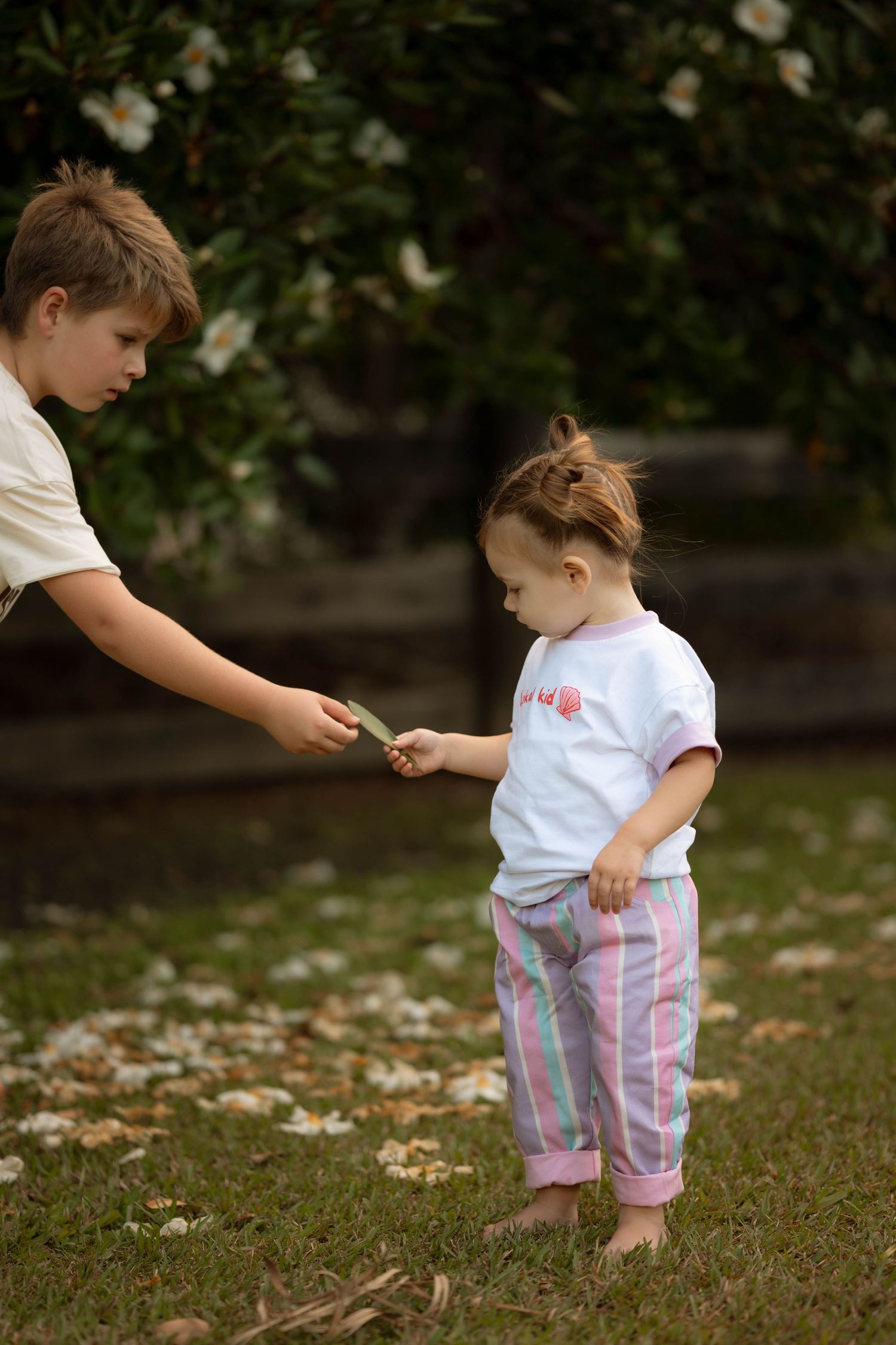 Boy handing a small flower to a toddler girl in pink striped pants outdoors.