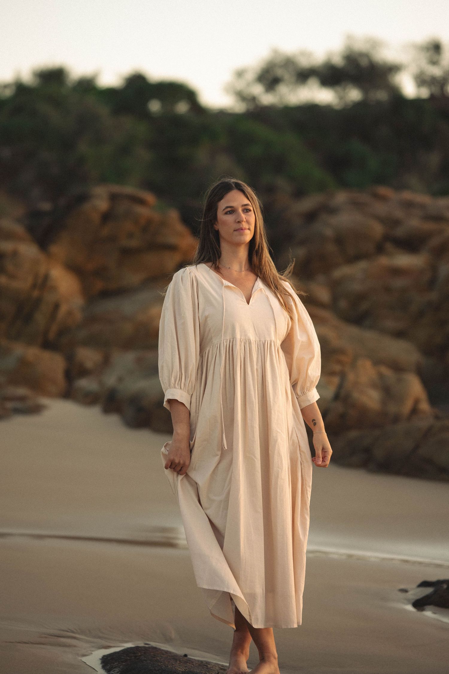 Woman in white maxi dress walking barefoot on sandy beach with rocky cliffs and trees in background.